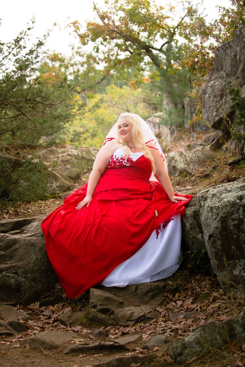 A flowing red ball gown dress creates a striking contrast against autumn leaves and rocky terrain in a woodland setting.