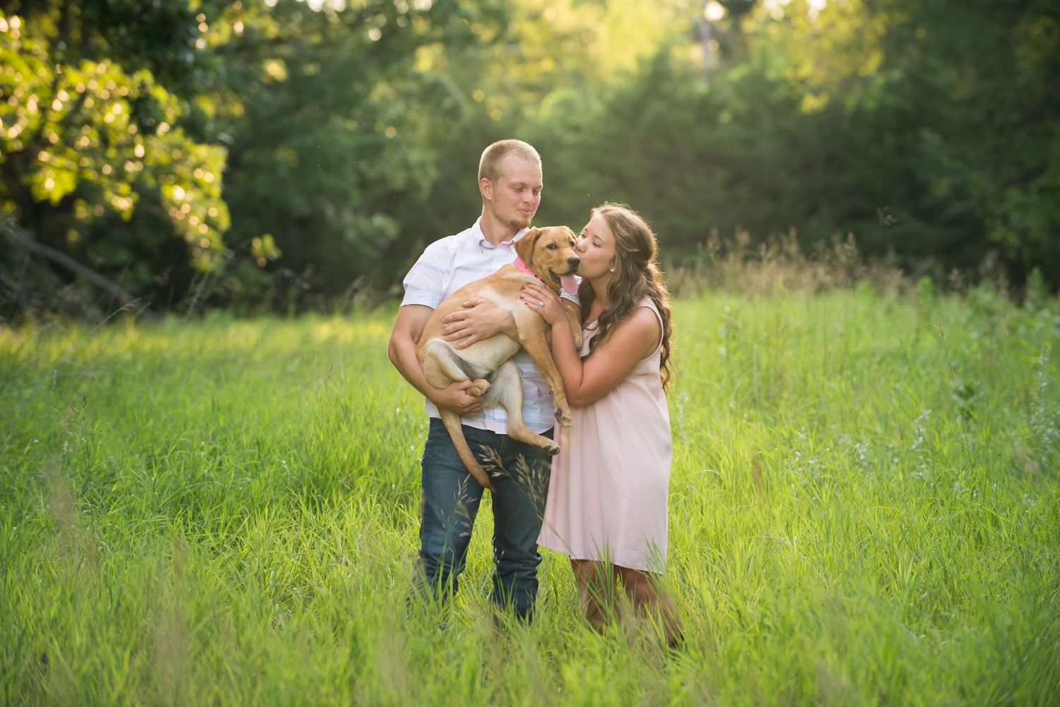 A couple shares a playful moment while standing in a sunlit meadow with golden light filtering through trees.