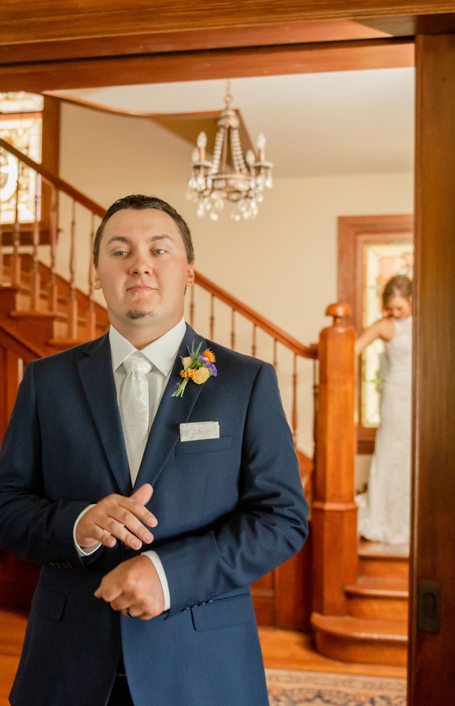 Person in formal navy suit with white tie and boutonniere standing indoors with wooden staircase visible.