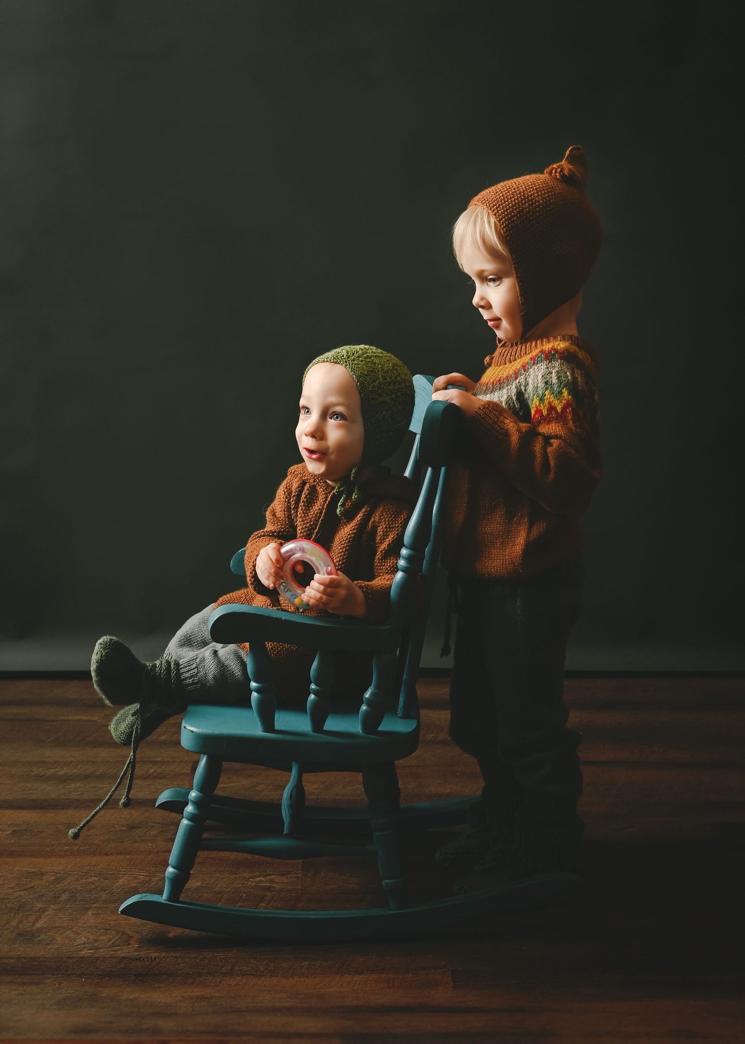 Two children interact around a blue wooden rocking chair against dark green background.