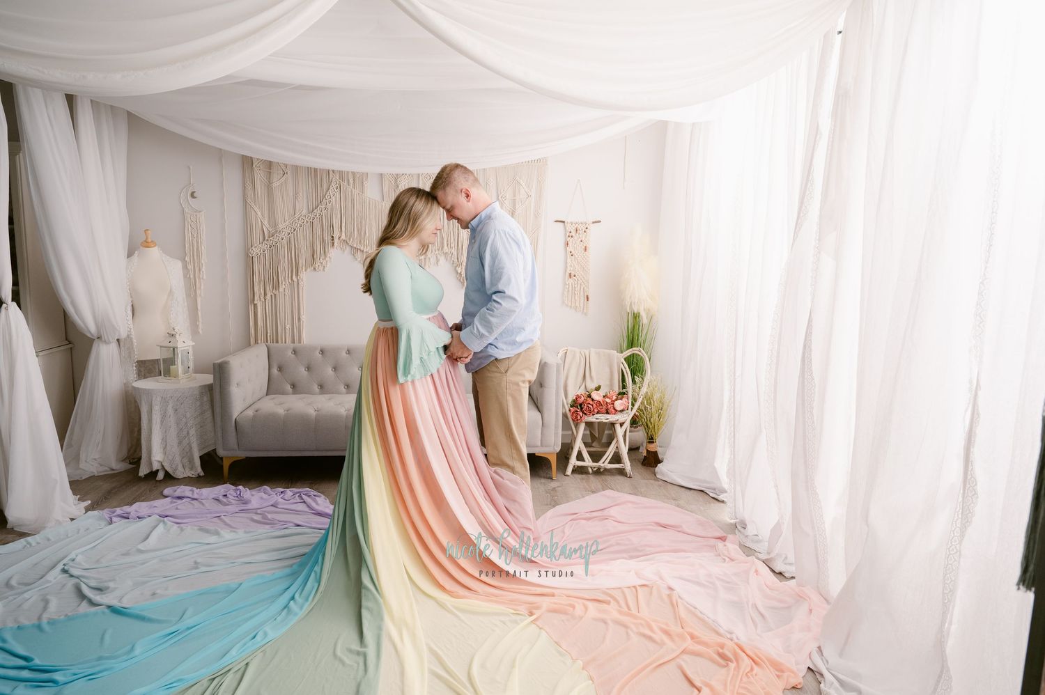 A couple embraces in a white draped room wearing pastel rainbow dress with flowing fabric on the floor.