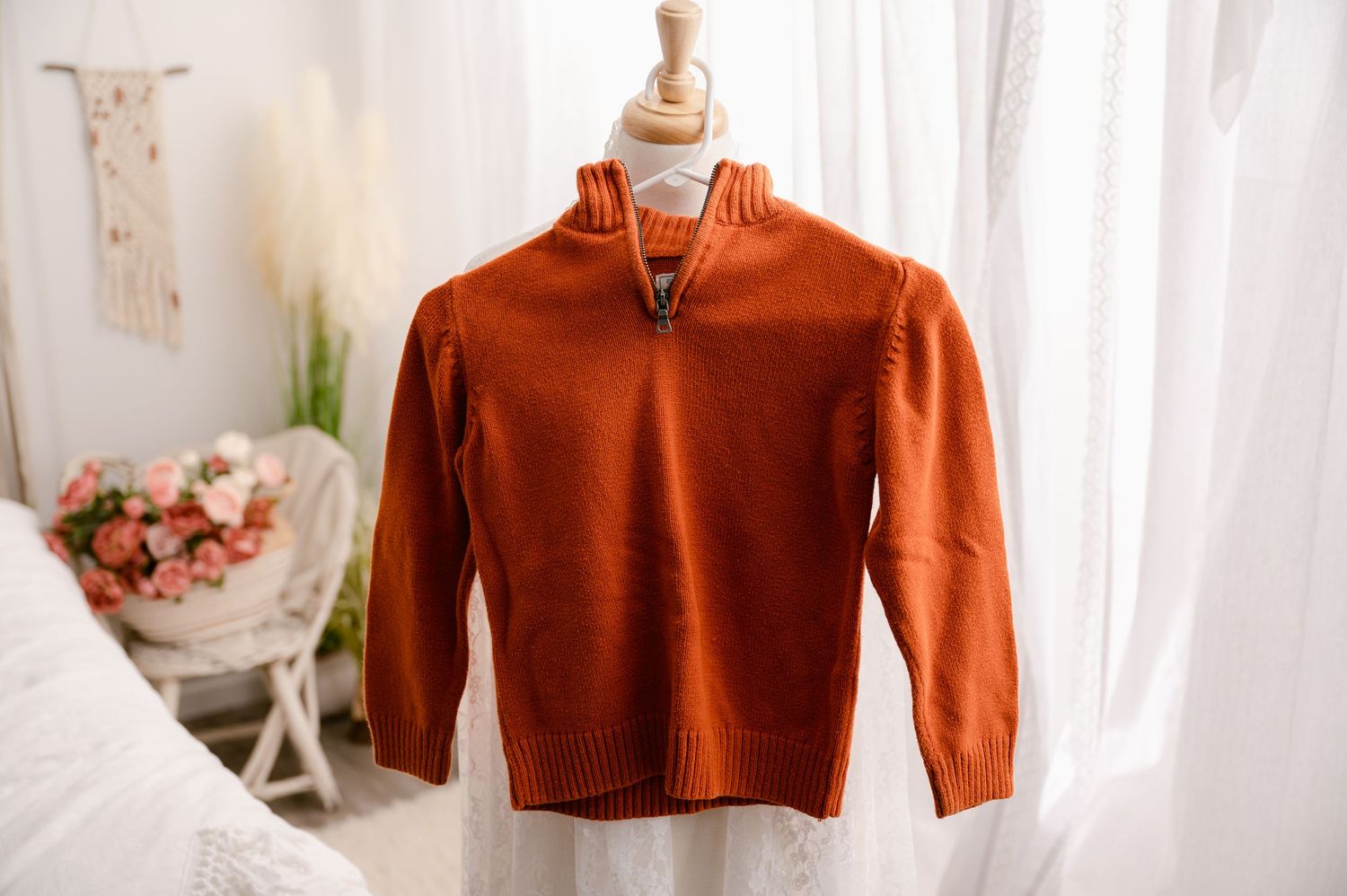 Burnt orange zip-up sweater displayed on wooden mannequin with soft natural lighting and floral decor in background.