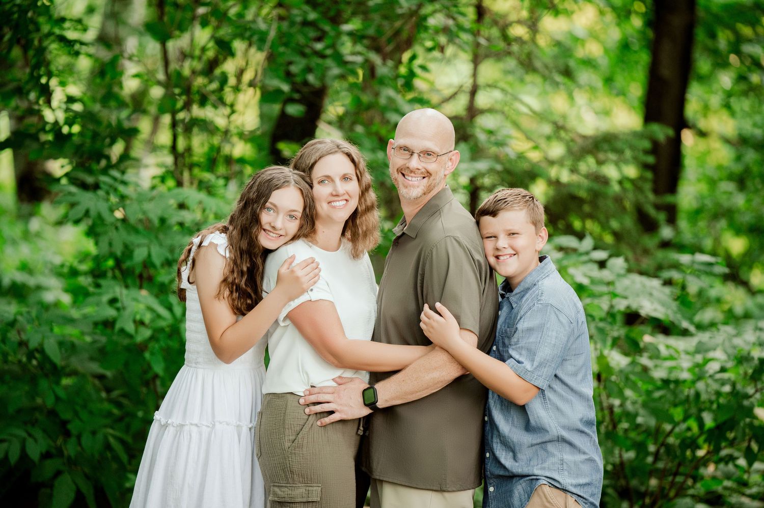Clear Lake MN Family Photos: A group embraces together while posing for a portrait in front of vibrant green forest foliage.
