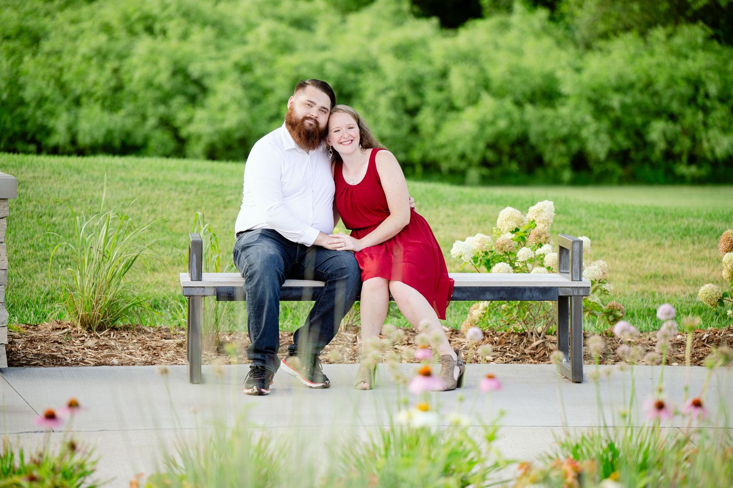 A couple sitting close together on a wooden bench in a lush garden setting with pink flowers blooming nearby.