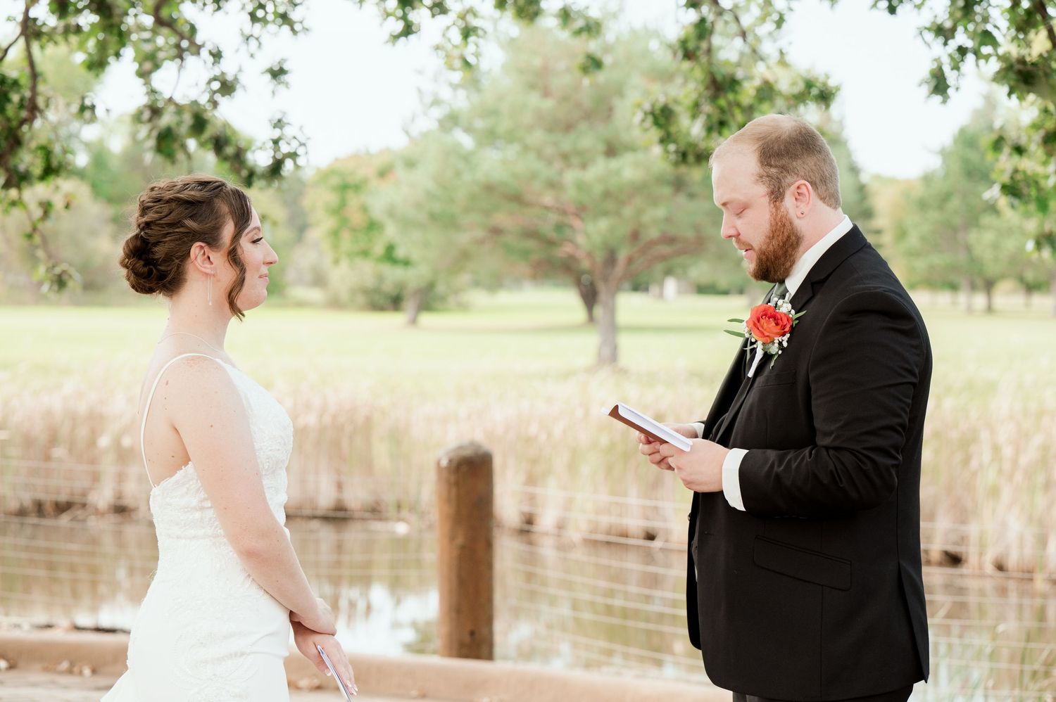 The Grands at Mulligans- Wedding Photos ceremony takes place outdoors by a lake with trees in the background as vows are exchanged.