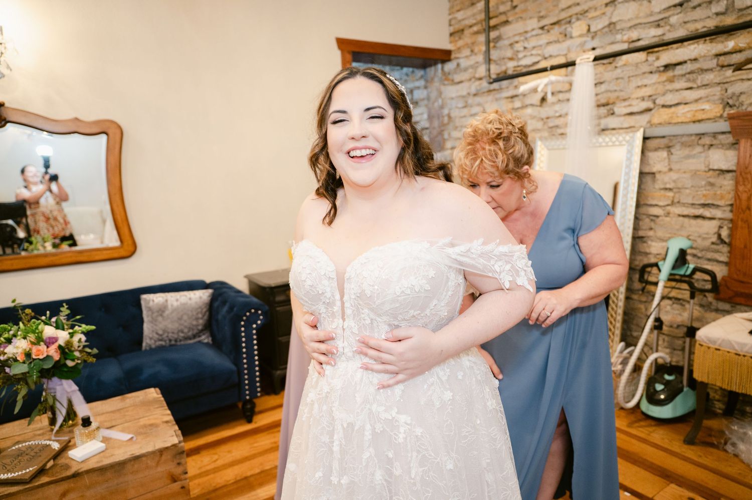 A joyful moment during wedding preparations shows the bride in an elegant off-shoulder lace gown against a rustic stone wall.