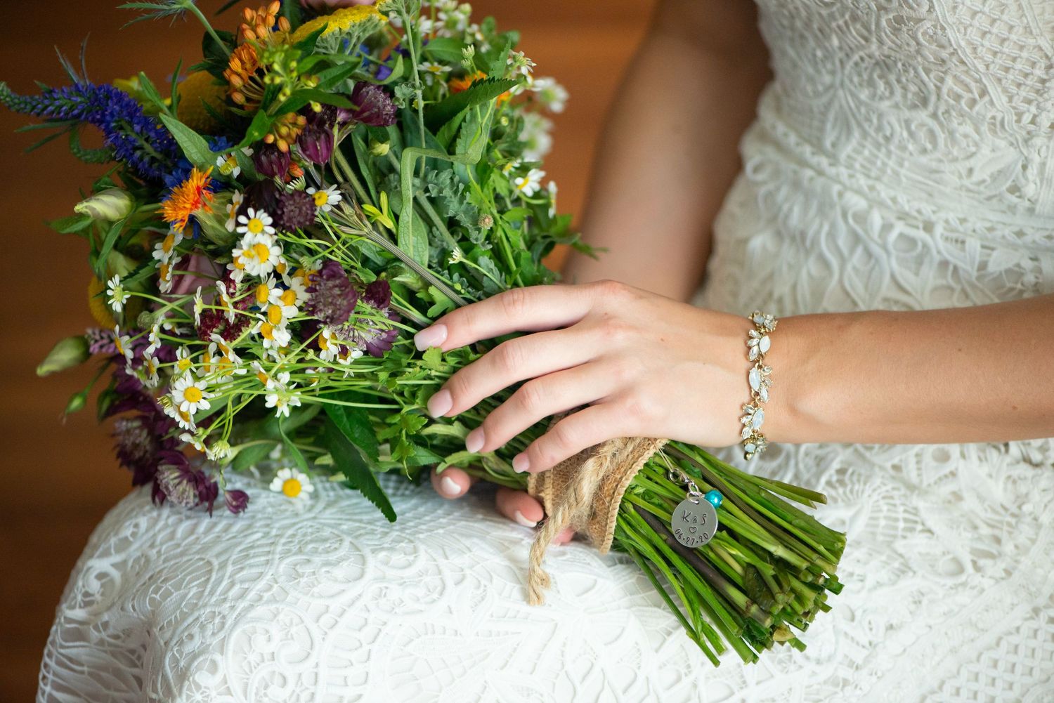 A wildflower wedding bouquet with purple, orange and yellow blooms is held against a white lace dress.