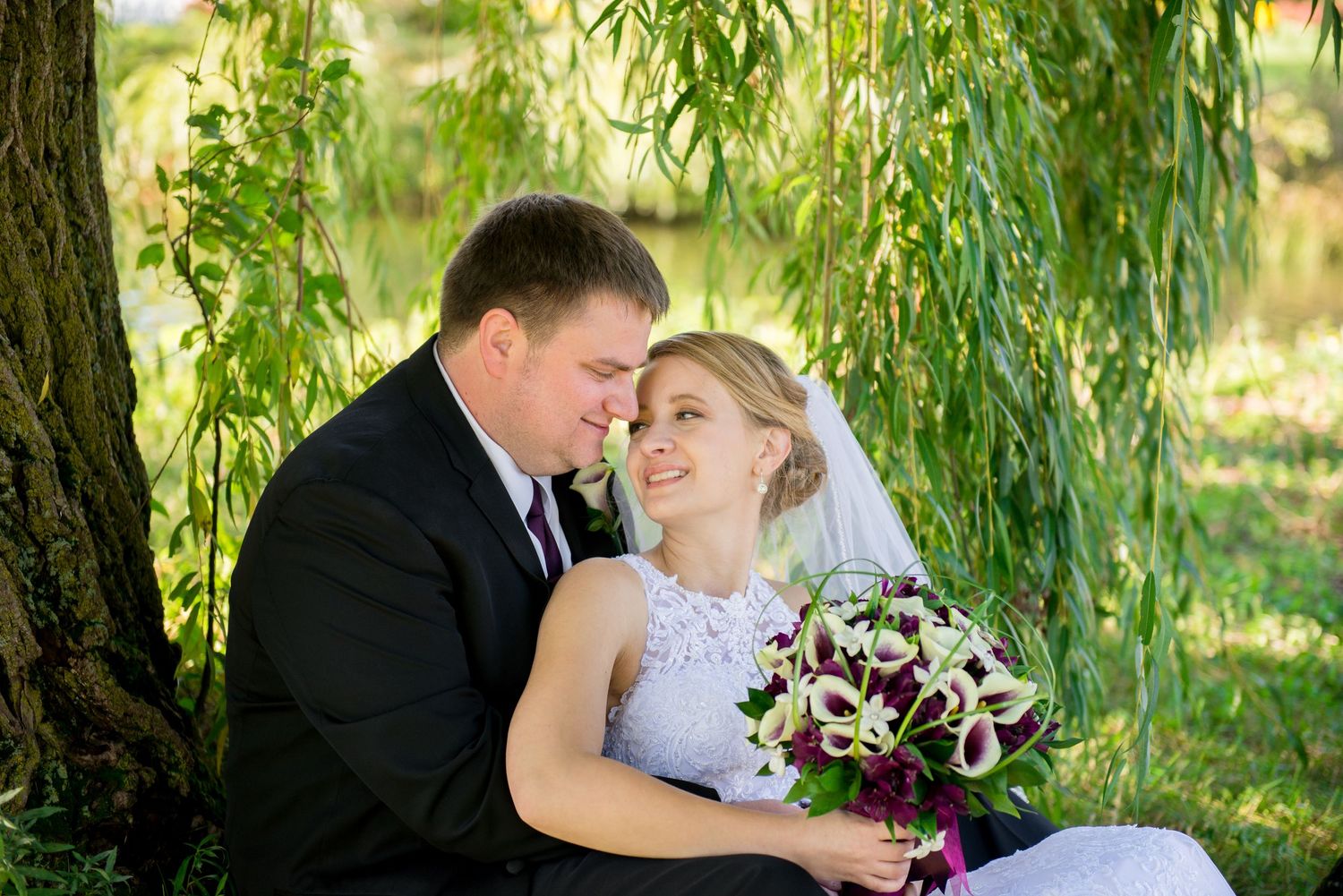 Couple shares tender moment under a weeping willow tree with a purple and white bouquet. Andover wedding at Magestic Oaks Golf Club
