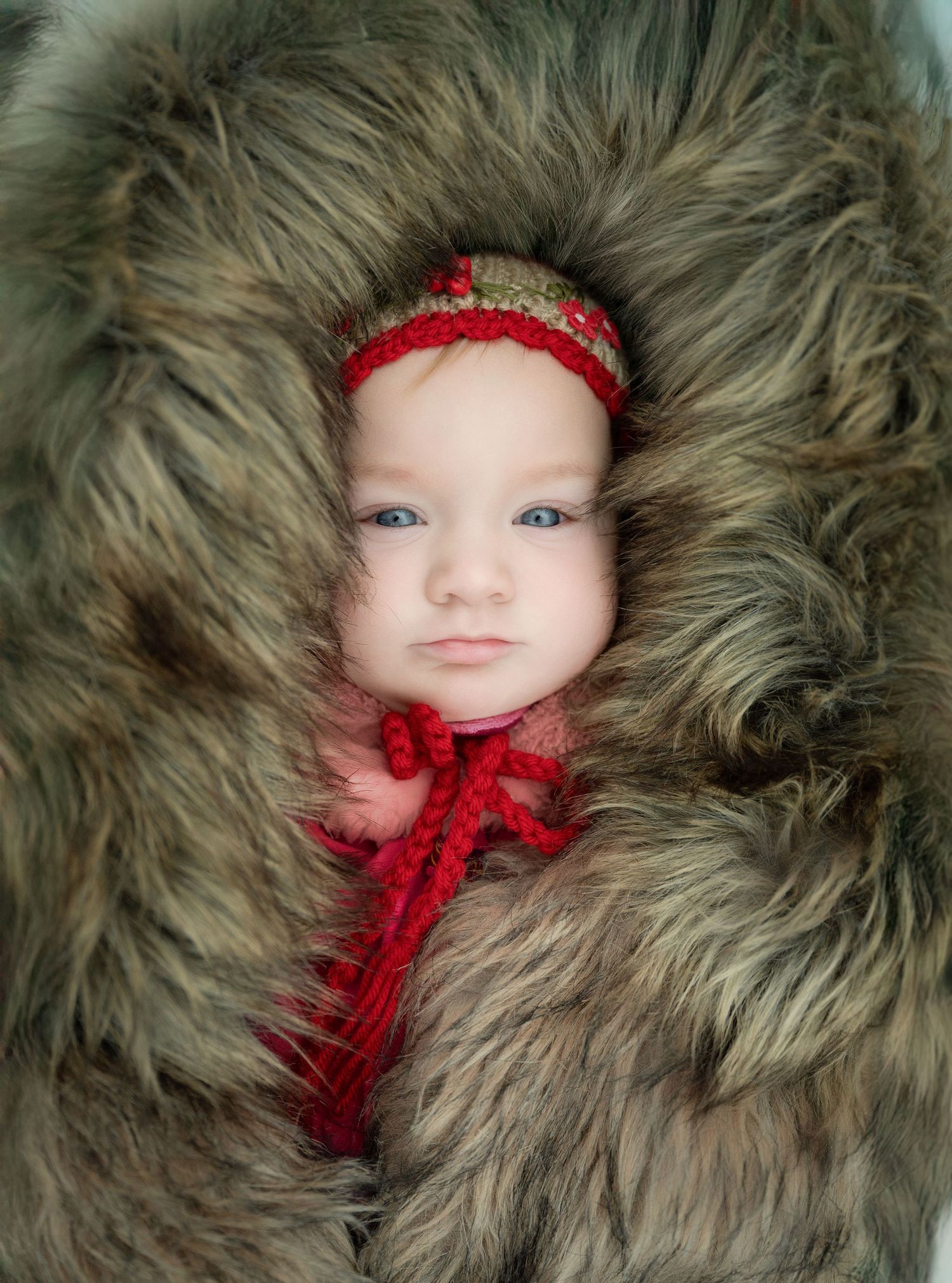 Small figure wearing a red ribbon nestled in soft gray furry fabric creating a cozy winter portrait setting.