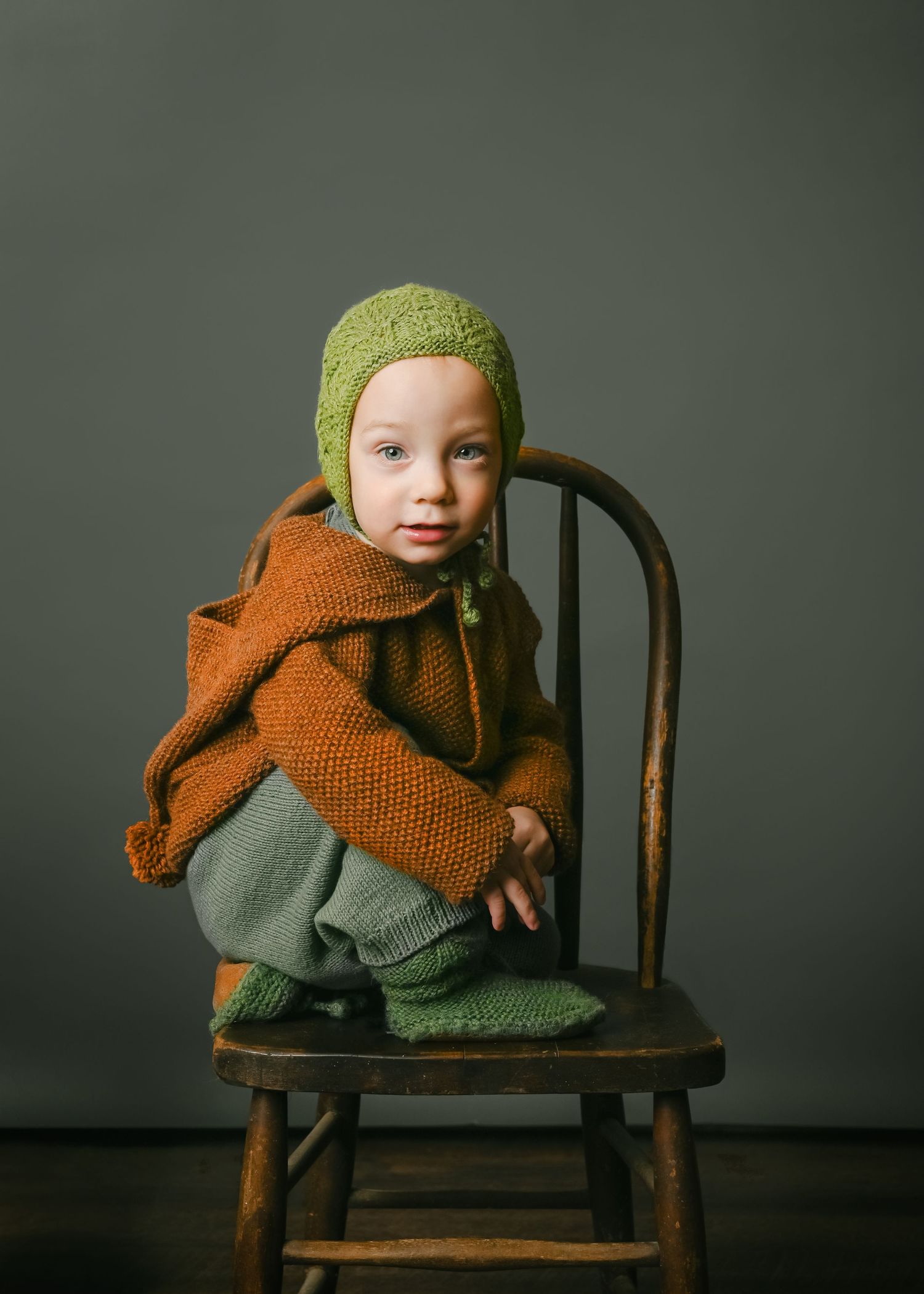 Child in brown sweater and green hat crouches on wooden chair against dark background.