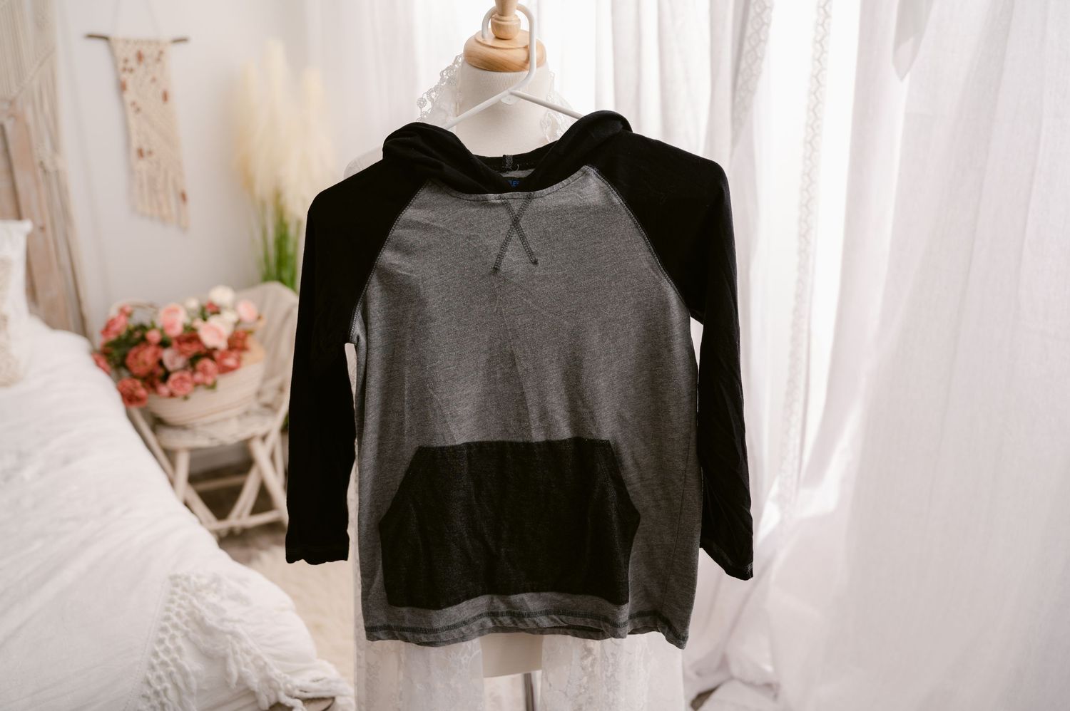A cozy black and gray raglan hoodie sweatshirt hangs against a white curtain backdrop with soft lighting.