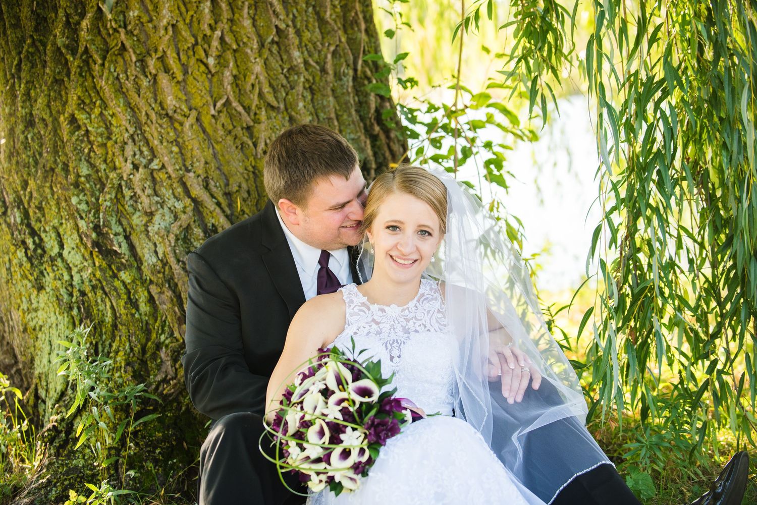 A wedding couple shares an intimate moment under a weeping willow tree with a purple bouquet. Andover wedding at Magestic Oaks Golf Club