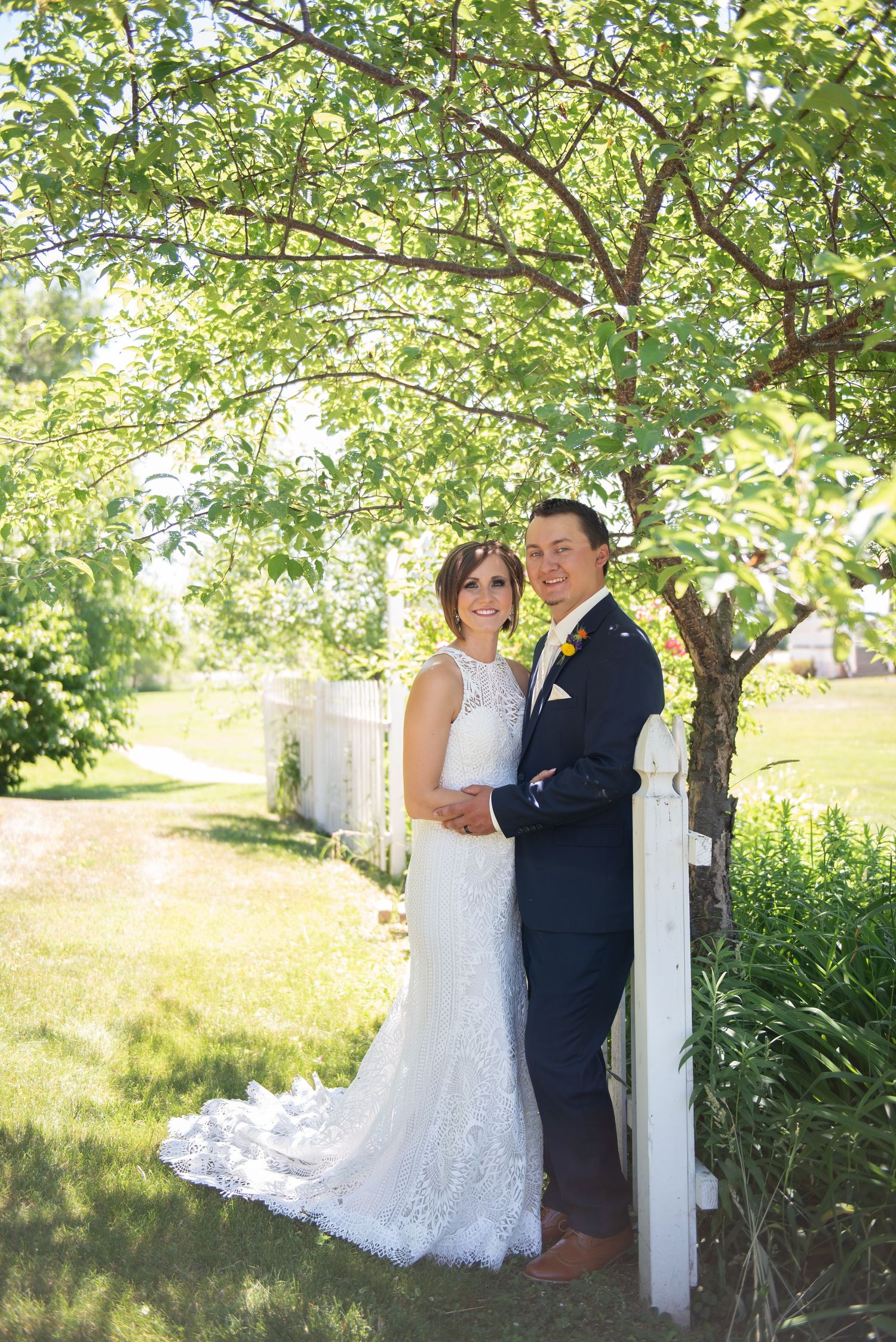 A wedding couple poses together by a white fence post under a leafy tree on a sunny summer day.