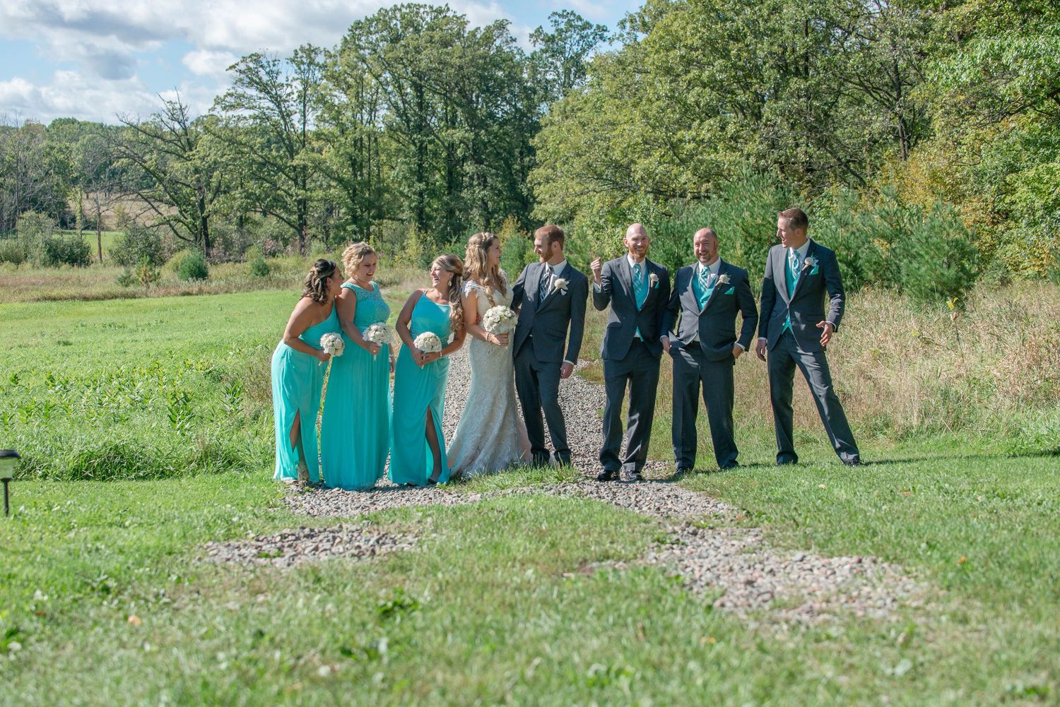 Wedding party in teal and navy attire poses together on a gravel path surrounded by lush green countryside. Rum River Barn Milaca Minnesota