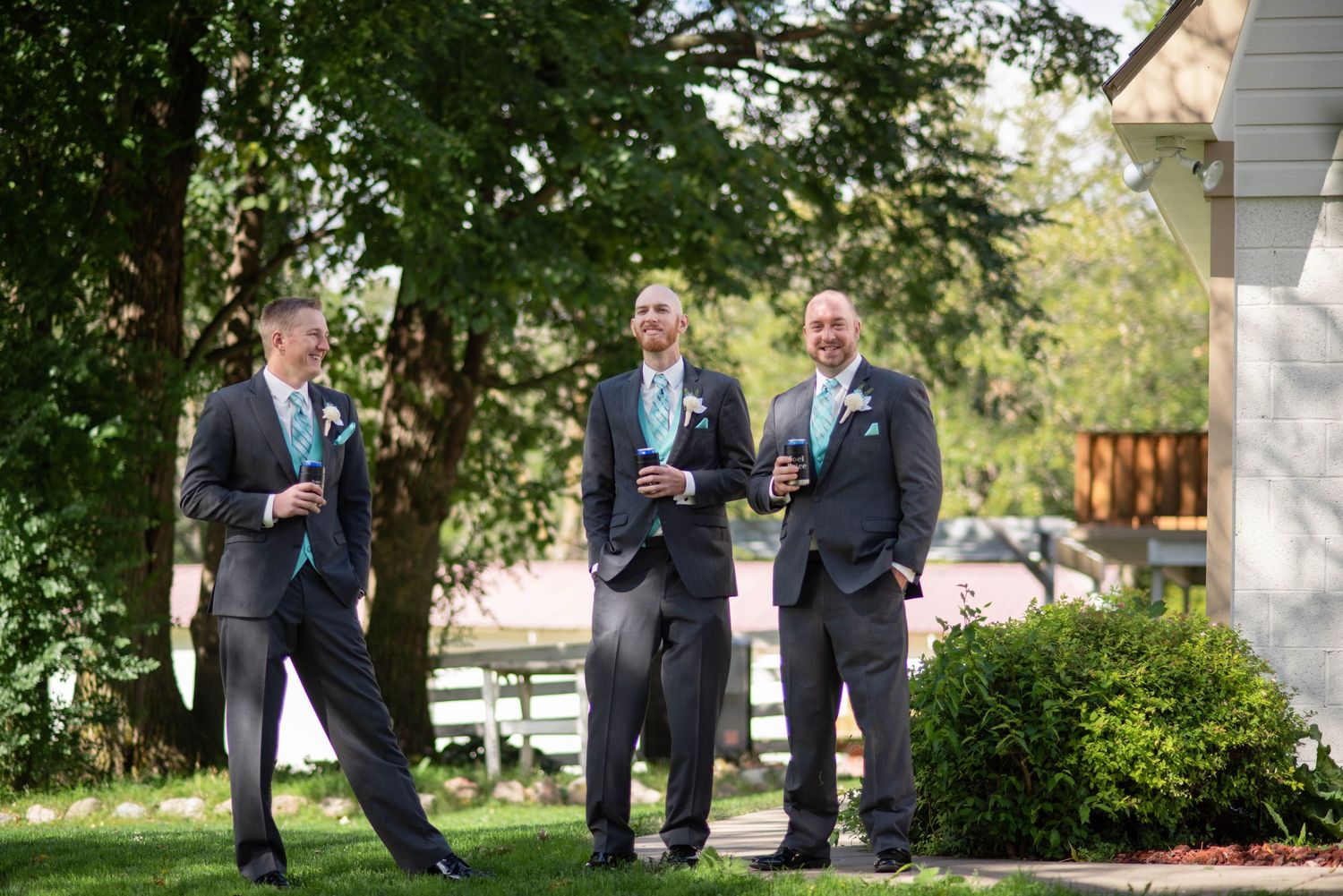 Three groomsmen in dark suits chat and laugh together in a garden setting before a wedding ceremony.