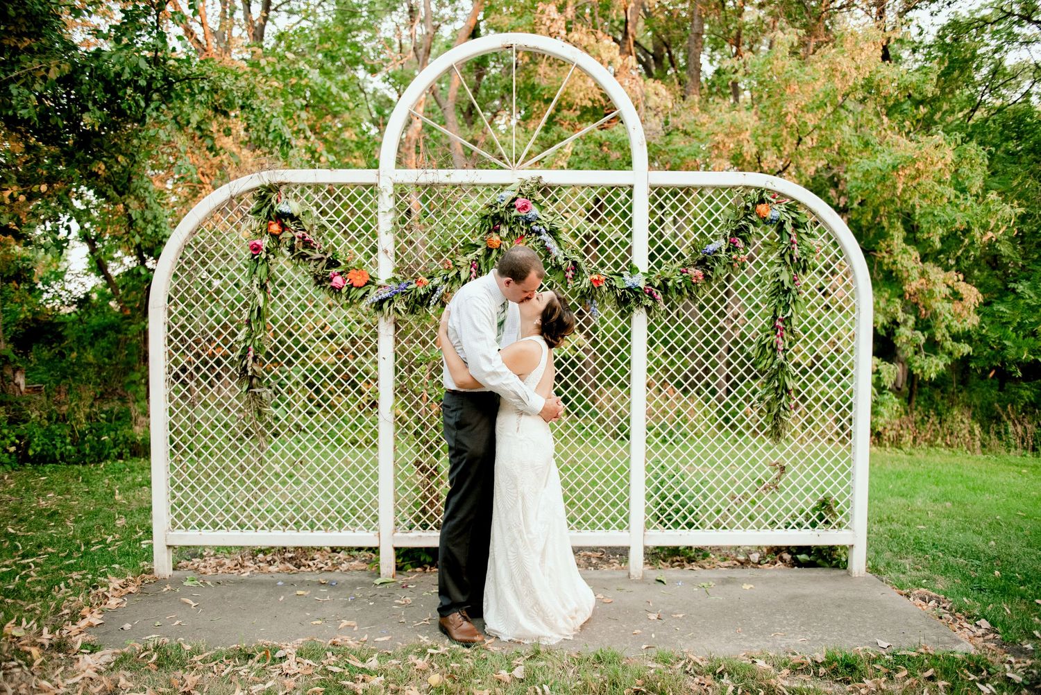 Romantic couple embraces under white lattice garden arch adorned with greenery in outdoor garden setting.