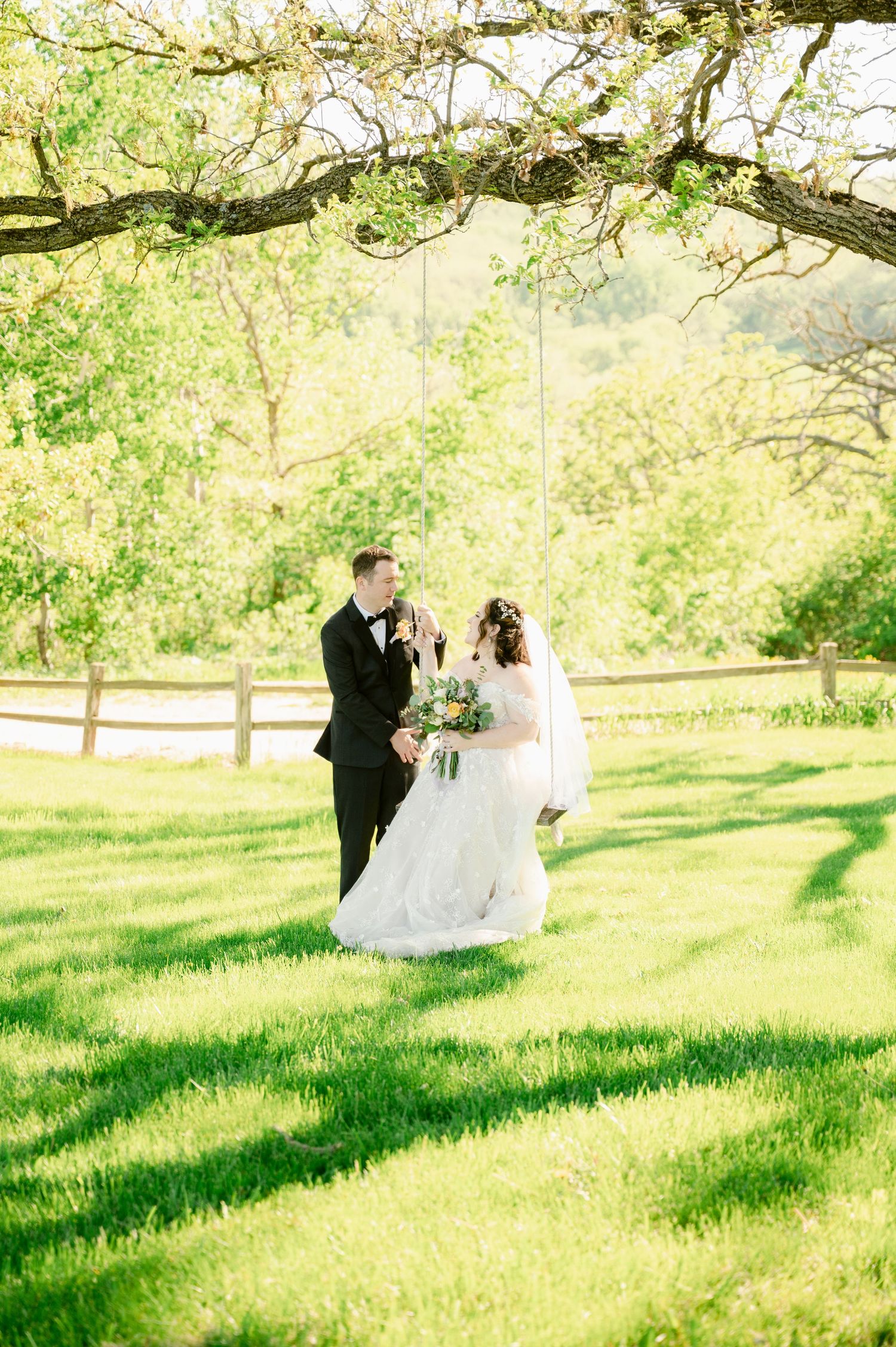 Wedding couple photographed under a tree arch in a sunlit meadow with wooden fencing.