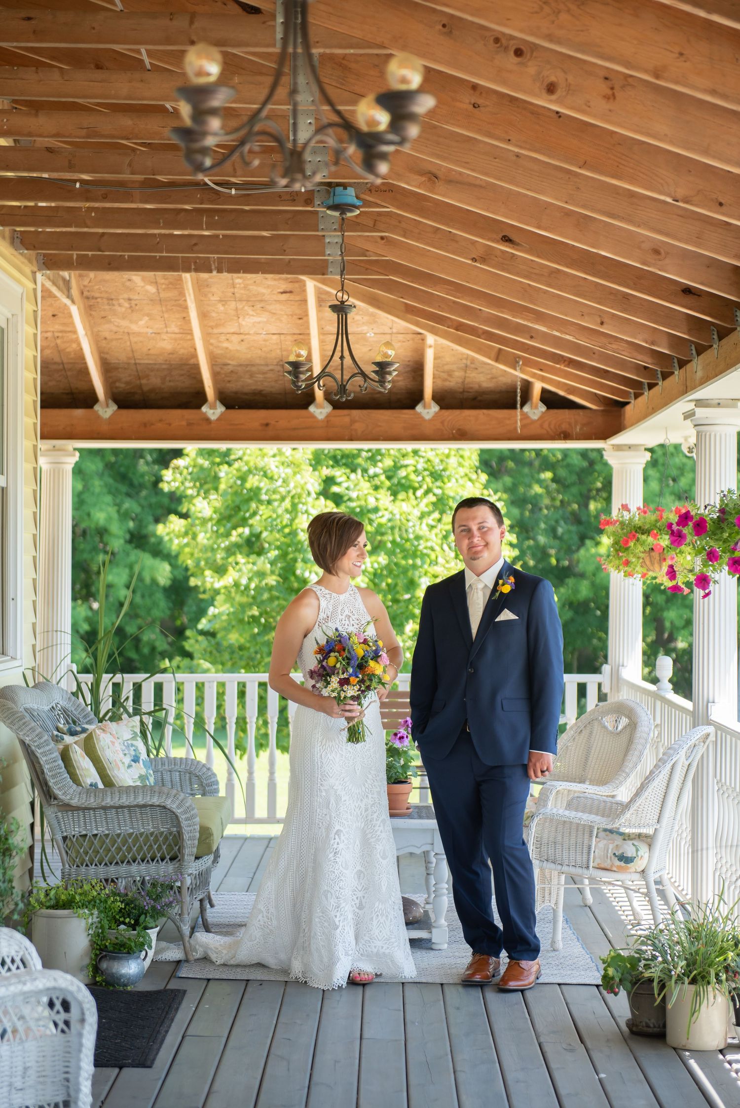 A couple poses for wedding photos on an elegant white porch with wicker furniture and hanging flower baskets.