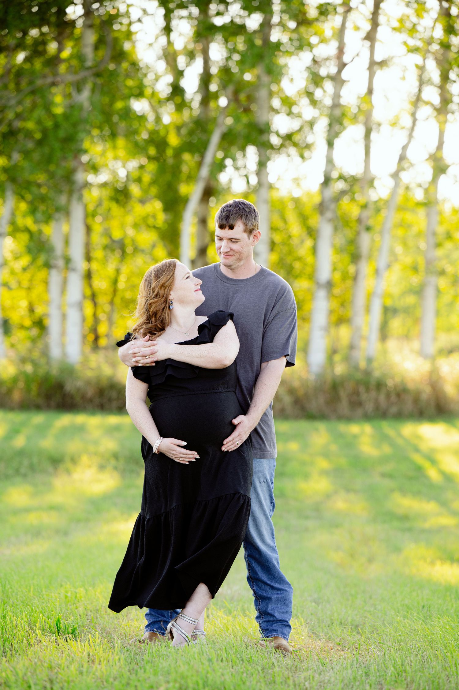 A couple embraces in casual clothing against a backdrop of birch trees during a maternity photo shoot.