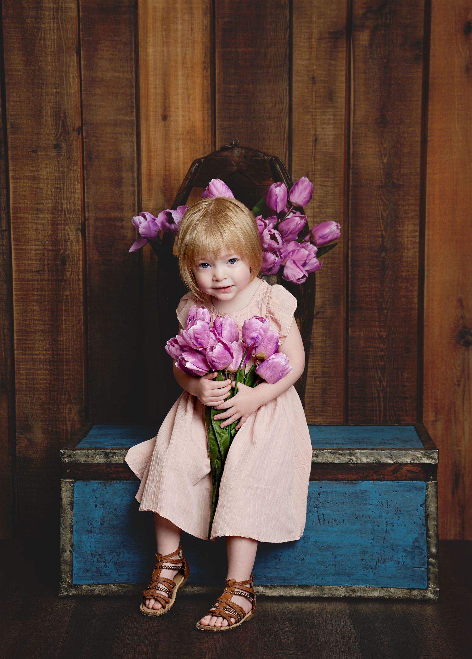 Young child in pink dress holding purple tulips while sitting on blue vintage chest.