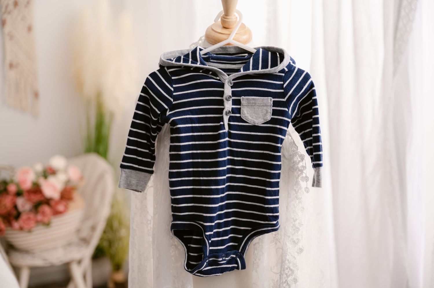 Navy and white striped baby bodysuit with grey pocket detail hanging on display against white lace curtain.