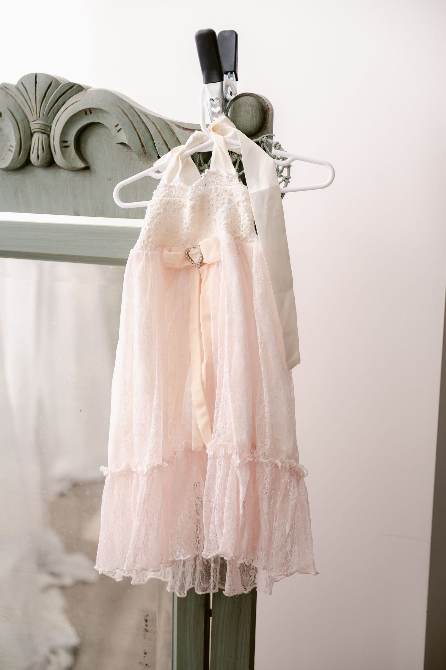 A delicate pink ruffled dress hangs on a white hanger against a vintage-style wall with ornate molding.