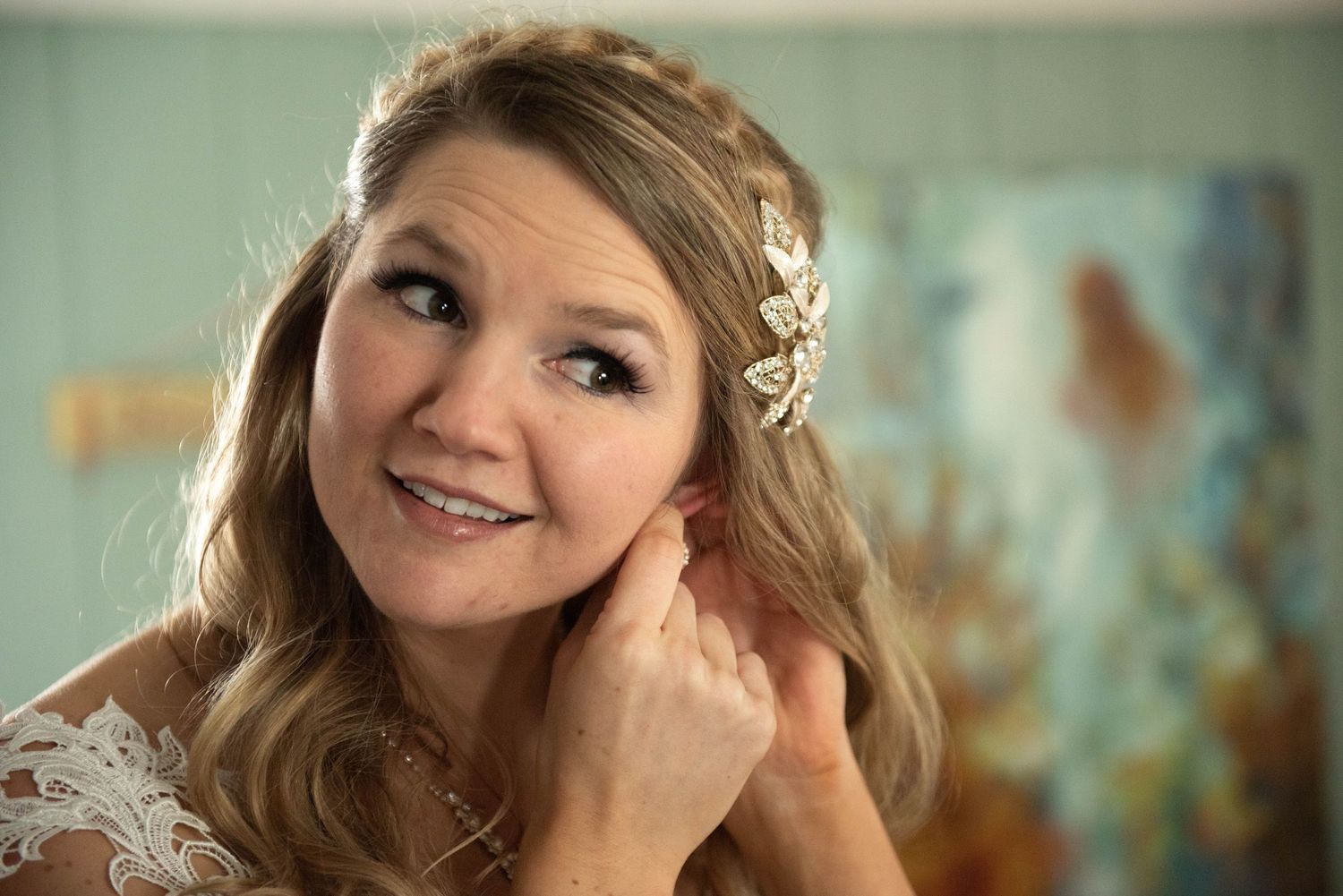 A bride wearing a white lace dress and delicate gold leaf headpiece smiles while touching her hair. Rum River Barn Milaca Minnesota Weddings