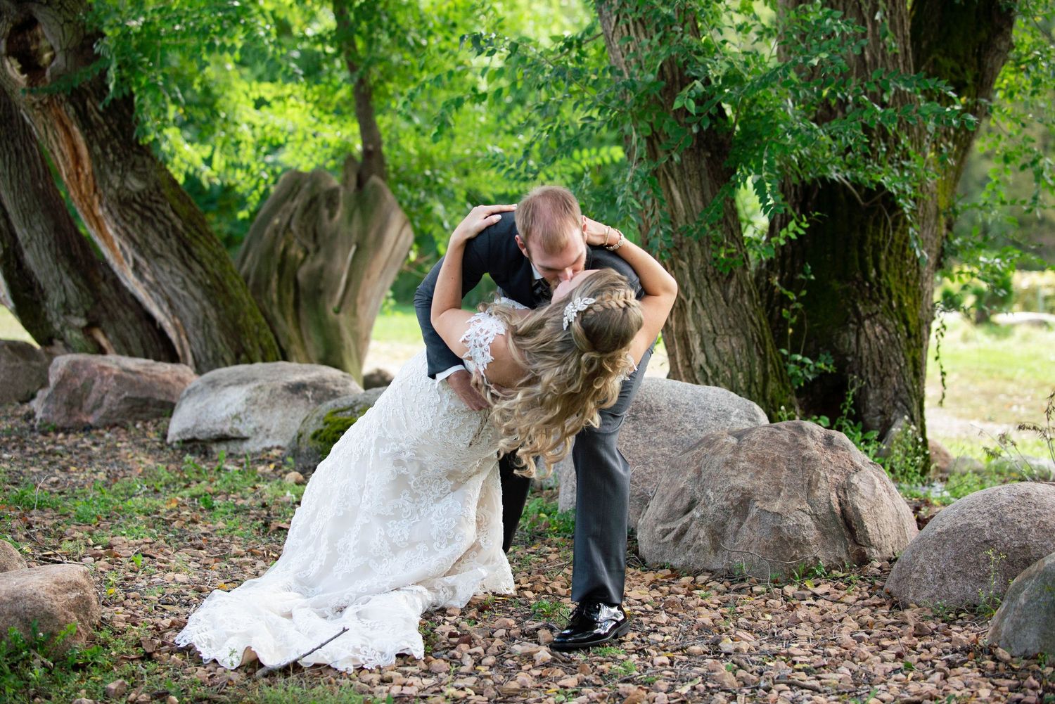 Romantic wedding portrait with couple embracing among trees and rocks in outdoor garden setting. Rum River Barn Milaca Minnesota Weddings
