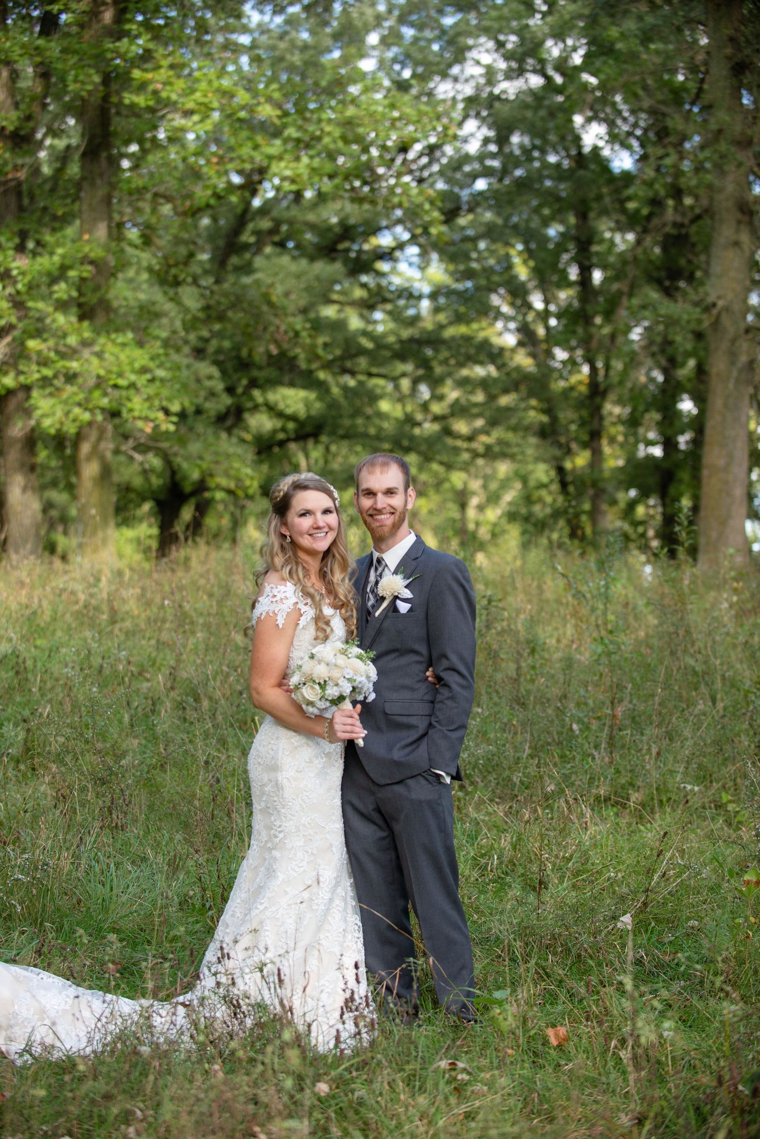 A wedding couple stands together in a sunlit forest clearing holding a white floral bouquet. Rum River Barn Milaca Minnesota Weddings