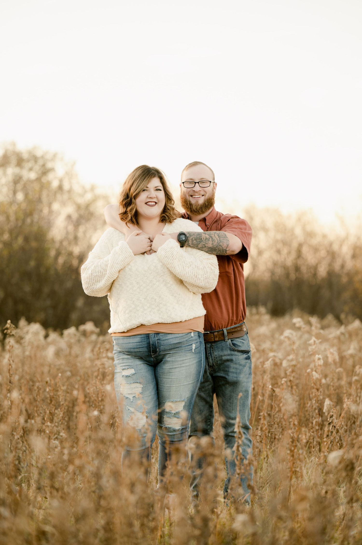 A couple stands together laughing in a golden sunlit field of tall grass during golden hour.
