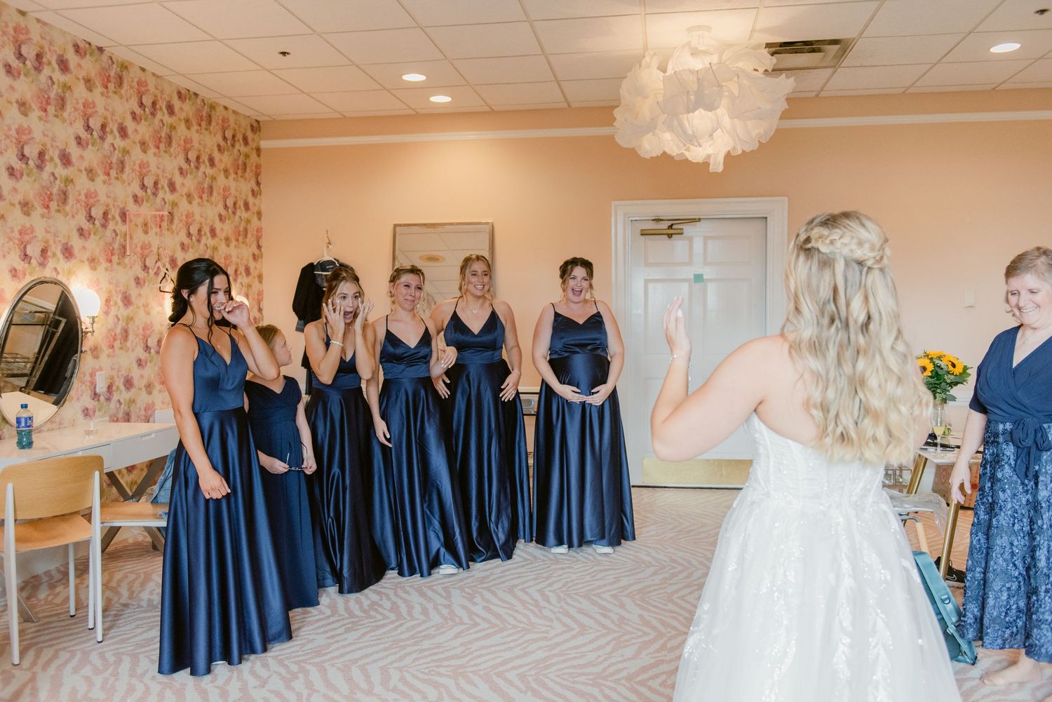Bridesmaids in matching navy blue gowns line up for a special reveal moment in a decorated room.