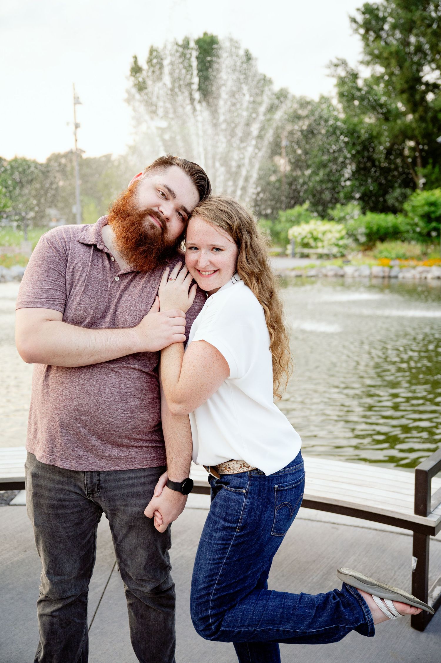 A playful couple poses together by a lake with decorative water fountains spraying behind them.