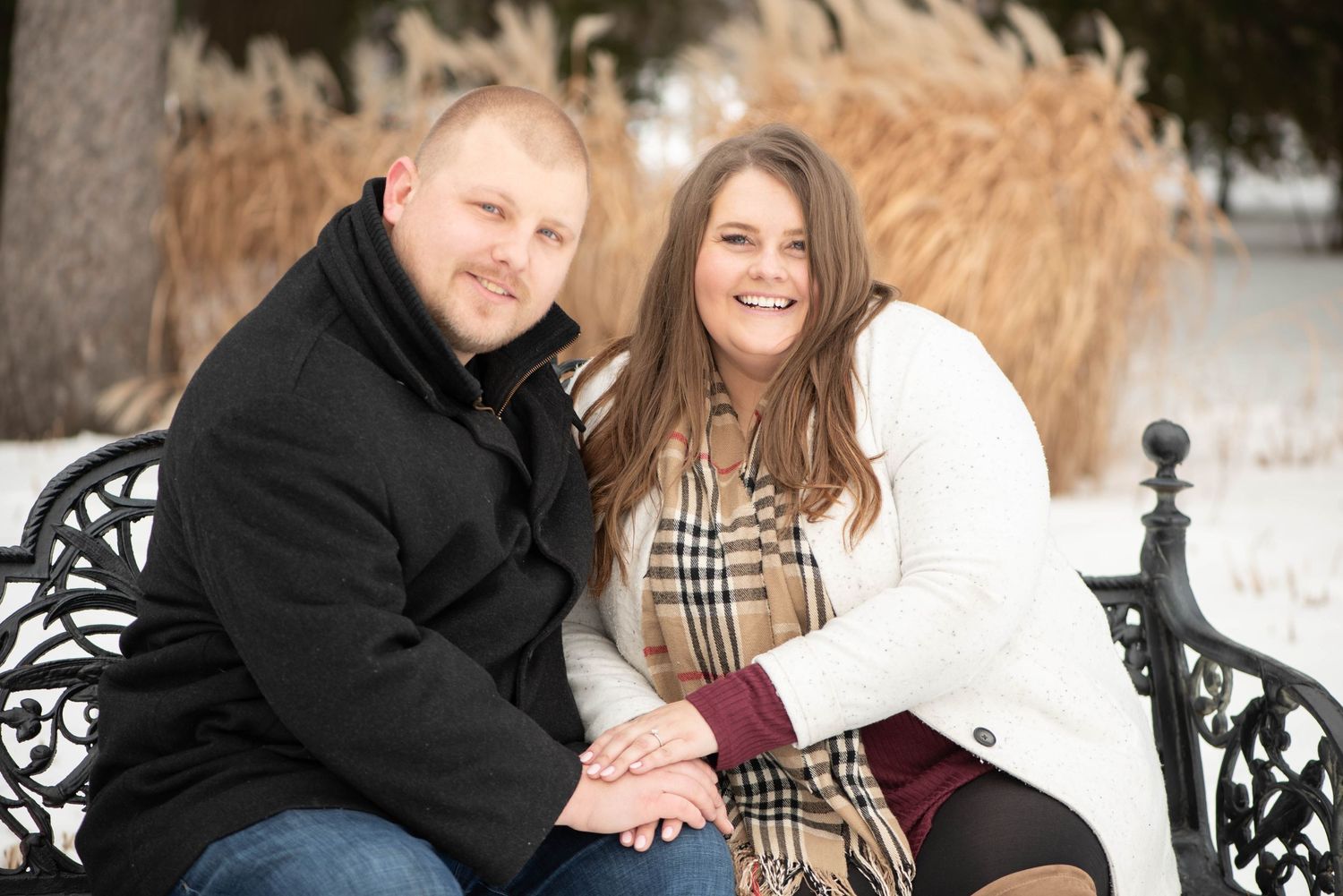 A couple sits together on an ornate black bench in a snowy winter scene with dried cattails in the background.
