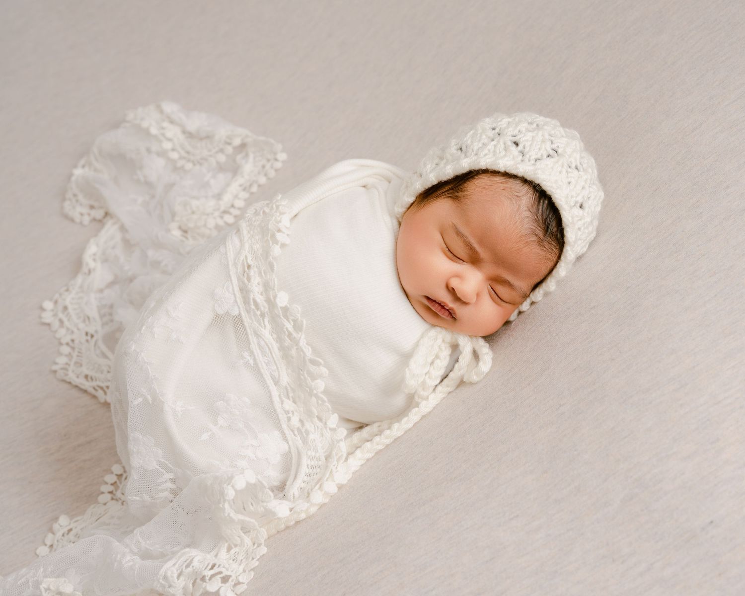 Sleeping newborn in vintage white lace christening gown and bonnet on neutral backdrop.