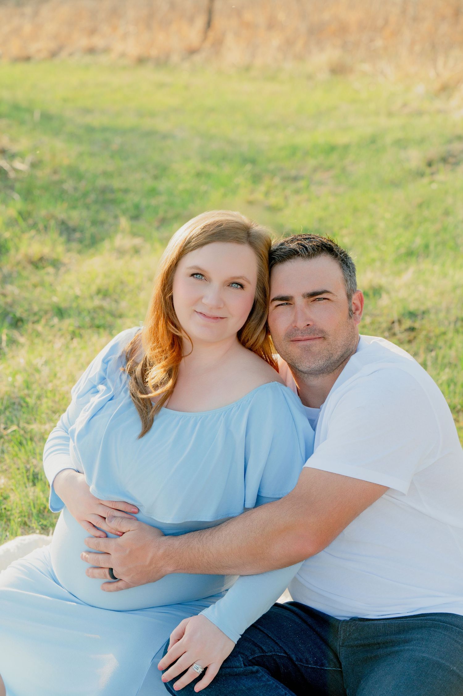 A couple embraces outdoors in a grassy field during a maternity photoshoot.