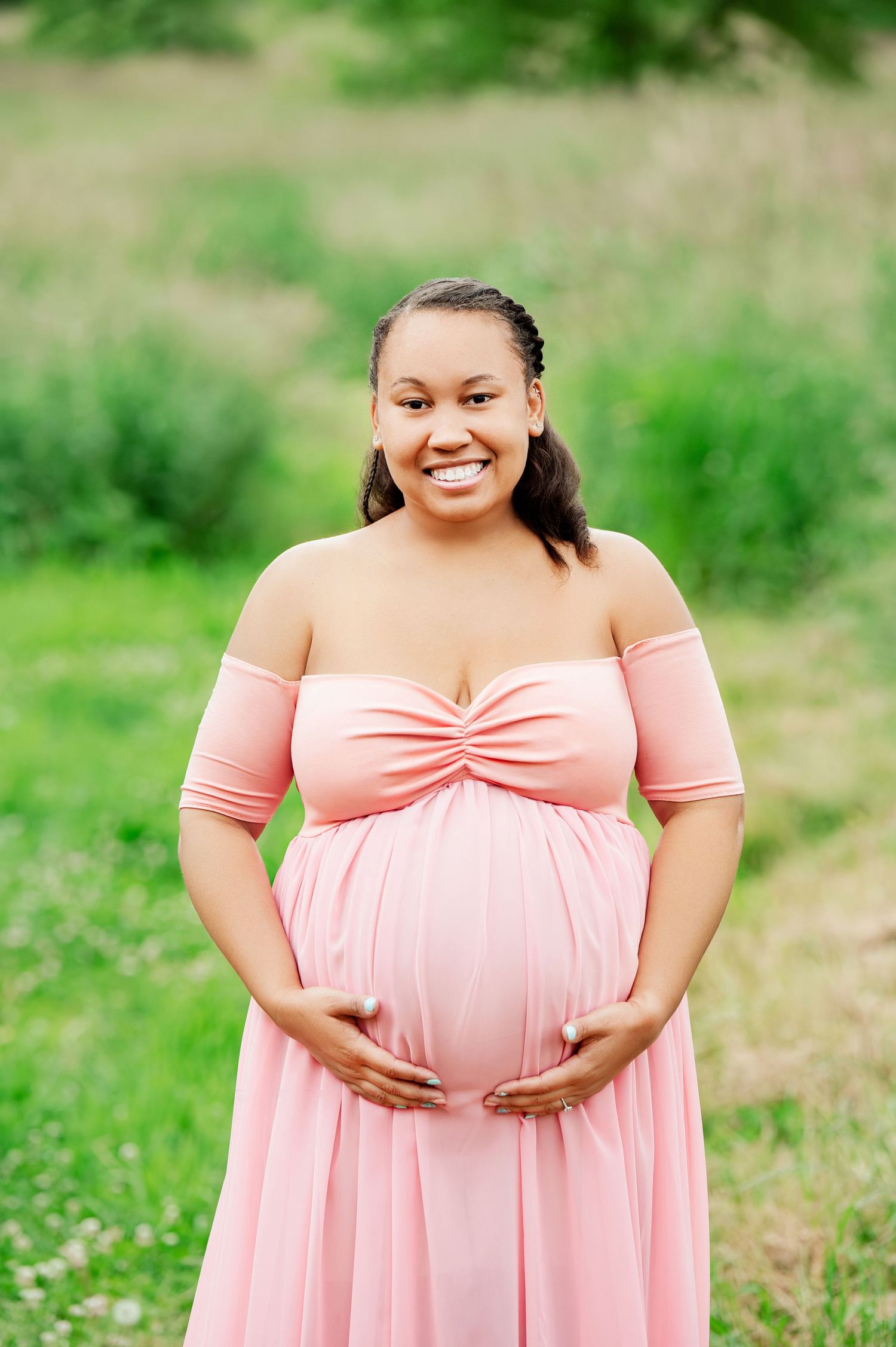 Pregnant woman in coral off-shoulder dress cradles belly while smiling in outdoor setting.