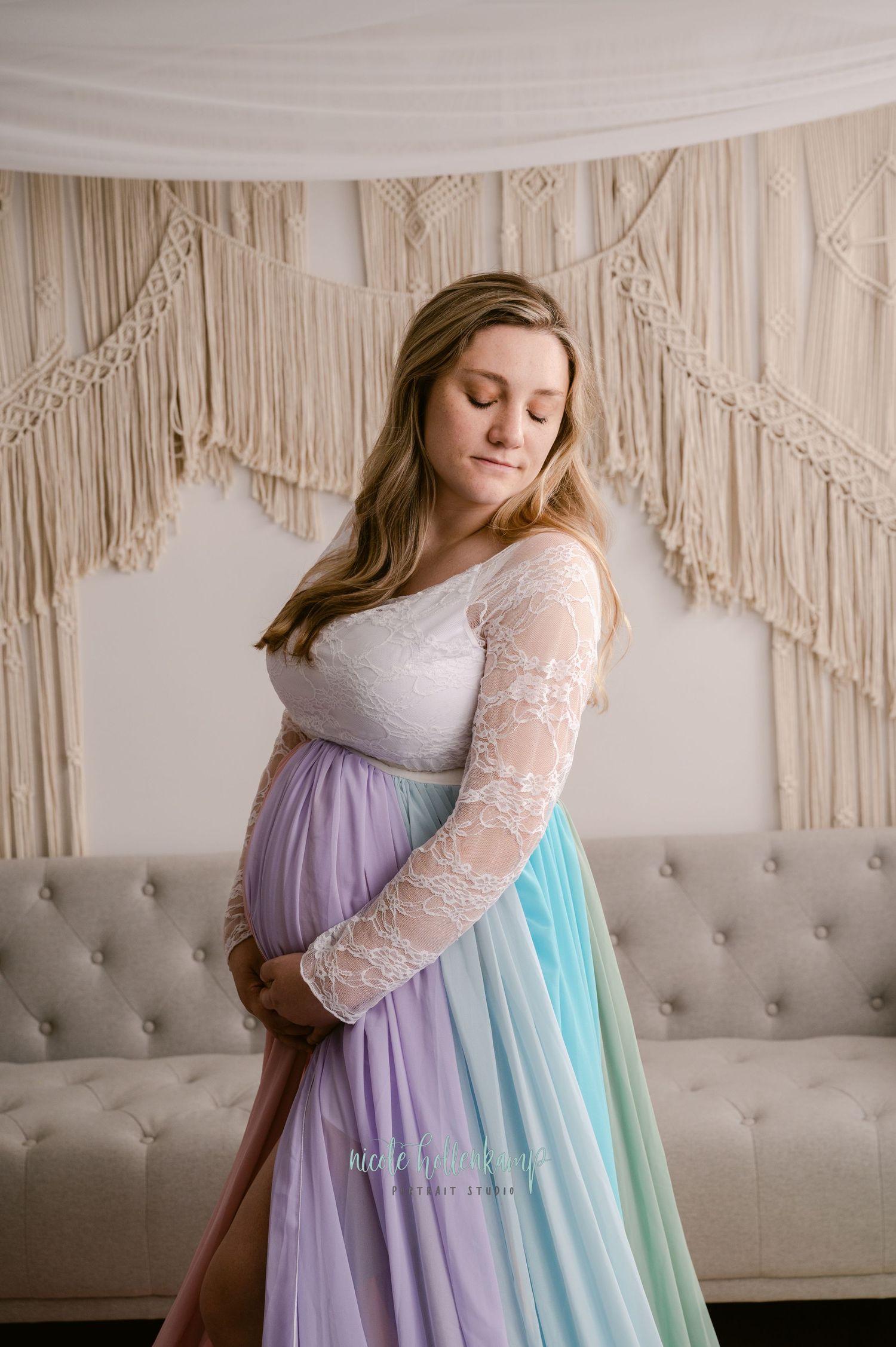 Expectant woman in lace sleeve dress poses against macrame backdrop wearing pastel rainbow maternity gown.