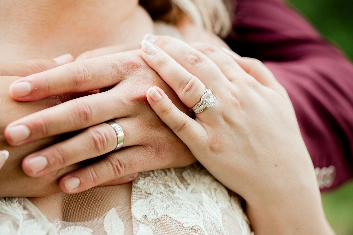 Close-up of hands displaying wedding rings against white lace dress and burgundy shirt in outdoor setting.