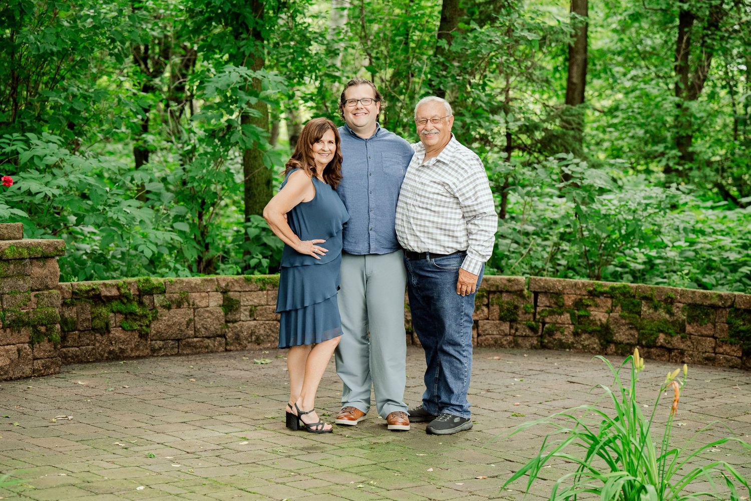 Clear Lake MN Family Photos: Three people pose for a family portrait on a stone patio surrounded by vibrant green forest.