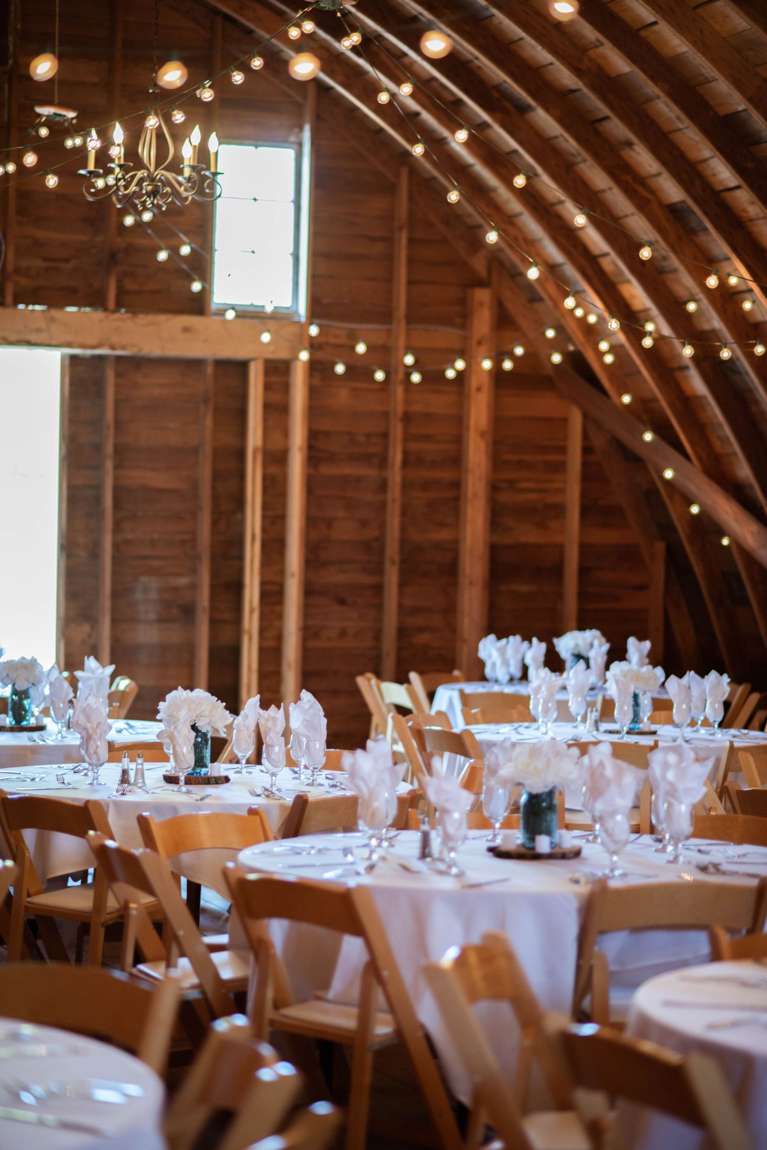 Rustic barn wedding reception setup with round tables, wooden chairs, and string lights under vaulted wooden ceiling.