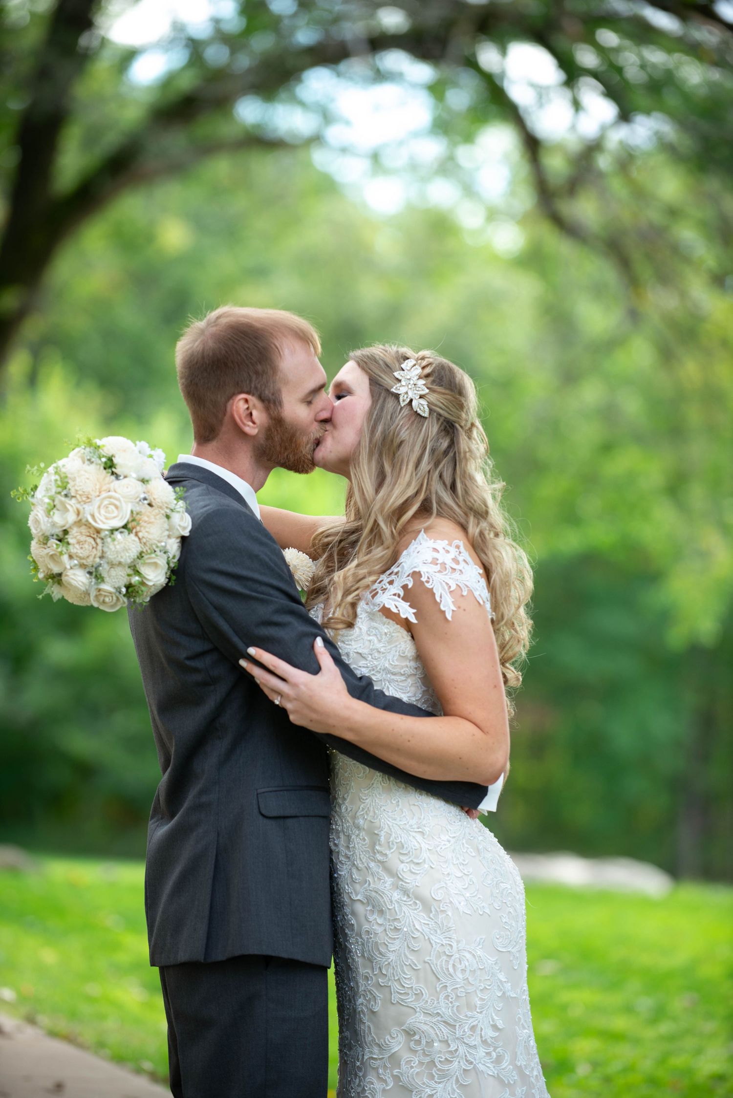A romantic wedding photo showing a couple kissing outdoors in a lush garden setting with a white floral bouquet. Rum River Barn Milaca Minne