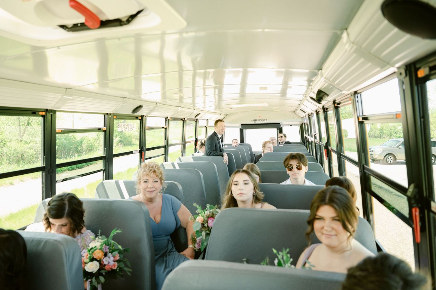 Wedding party members sit scattered throughout a school bus, holding bouquets and dressed in formal attire.