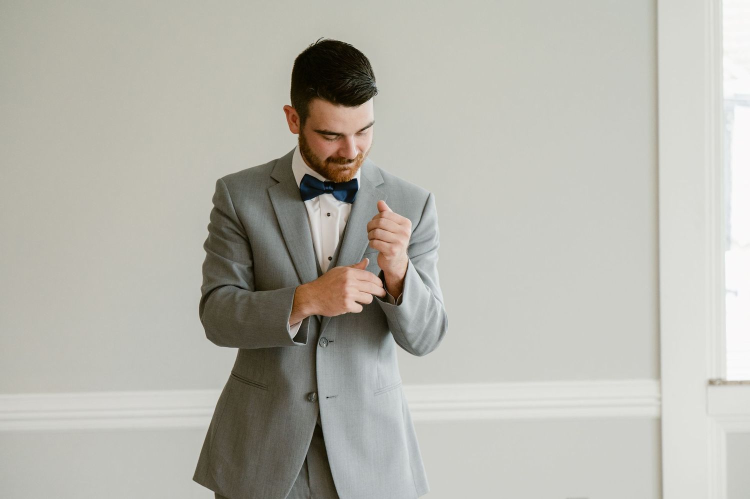 A formal portrait of someone in a gray suit and bowtie adjusting their cufflinks.