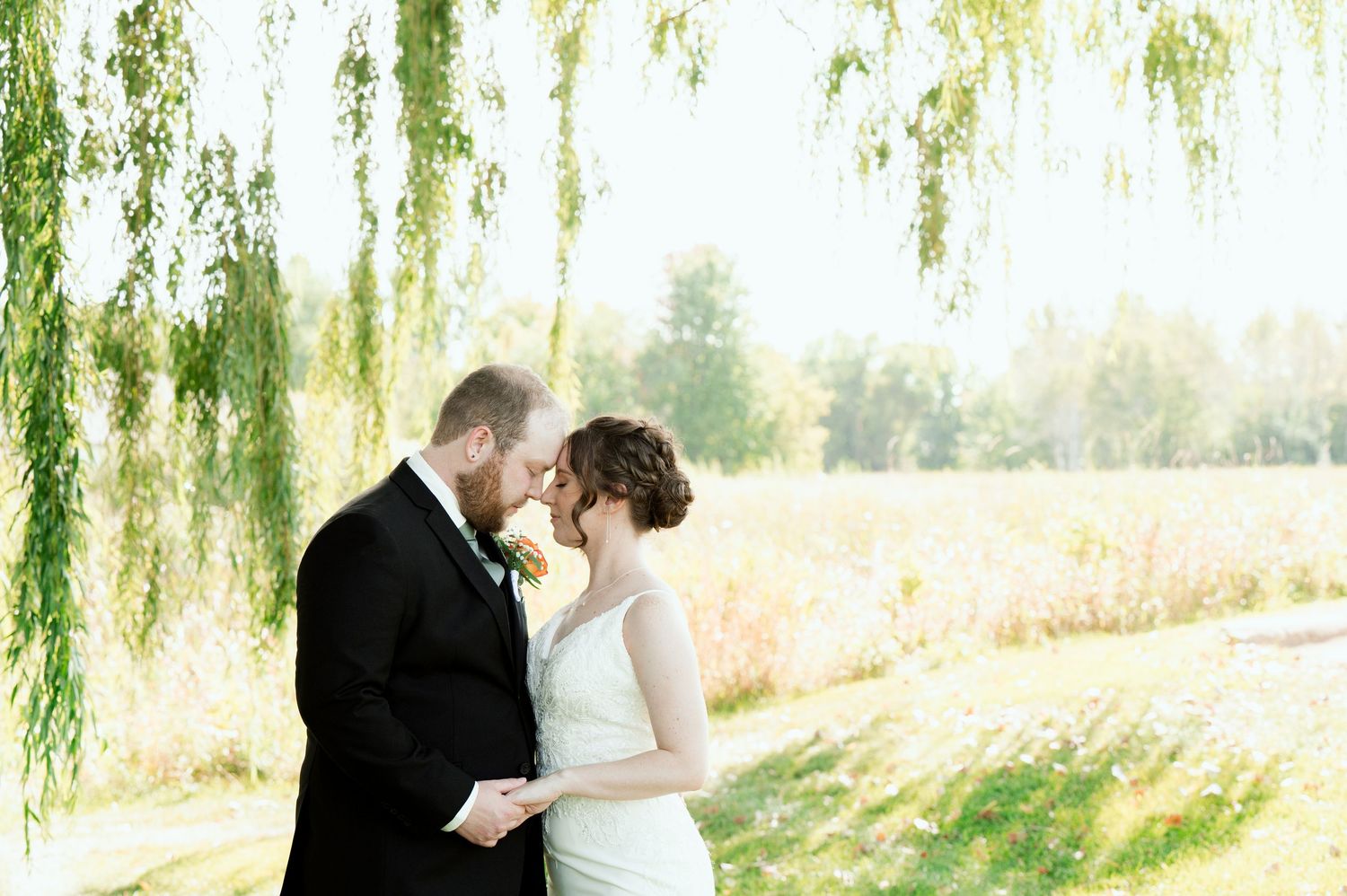 The Grands At Mulligans. Couple in formal attire shares an intimate moment under a cascading willow tree in golden sunlight.