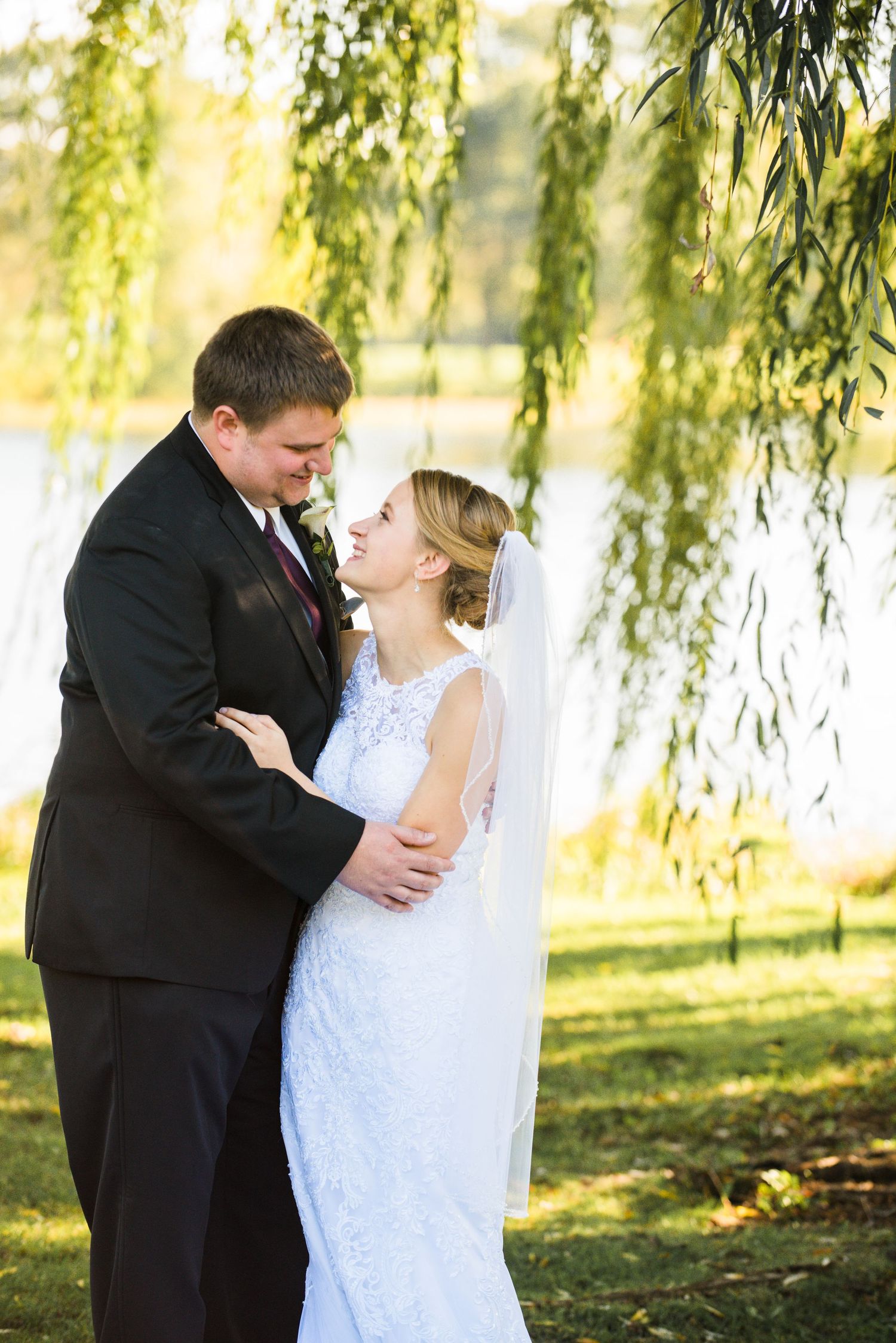 Joyful wedding couple sharing an intimate moment beneath willow trees by a lake. Andover wedding at Magestic Oaks Golf Club