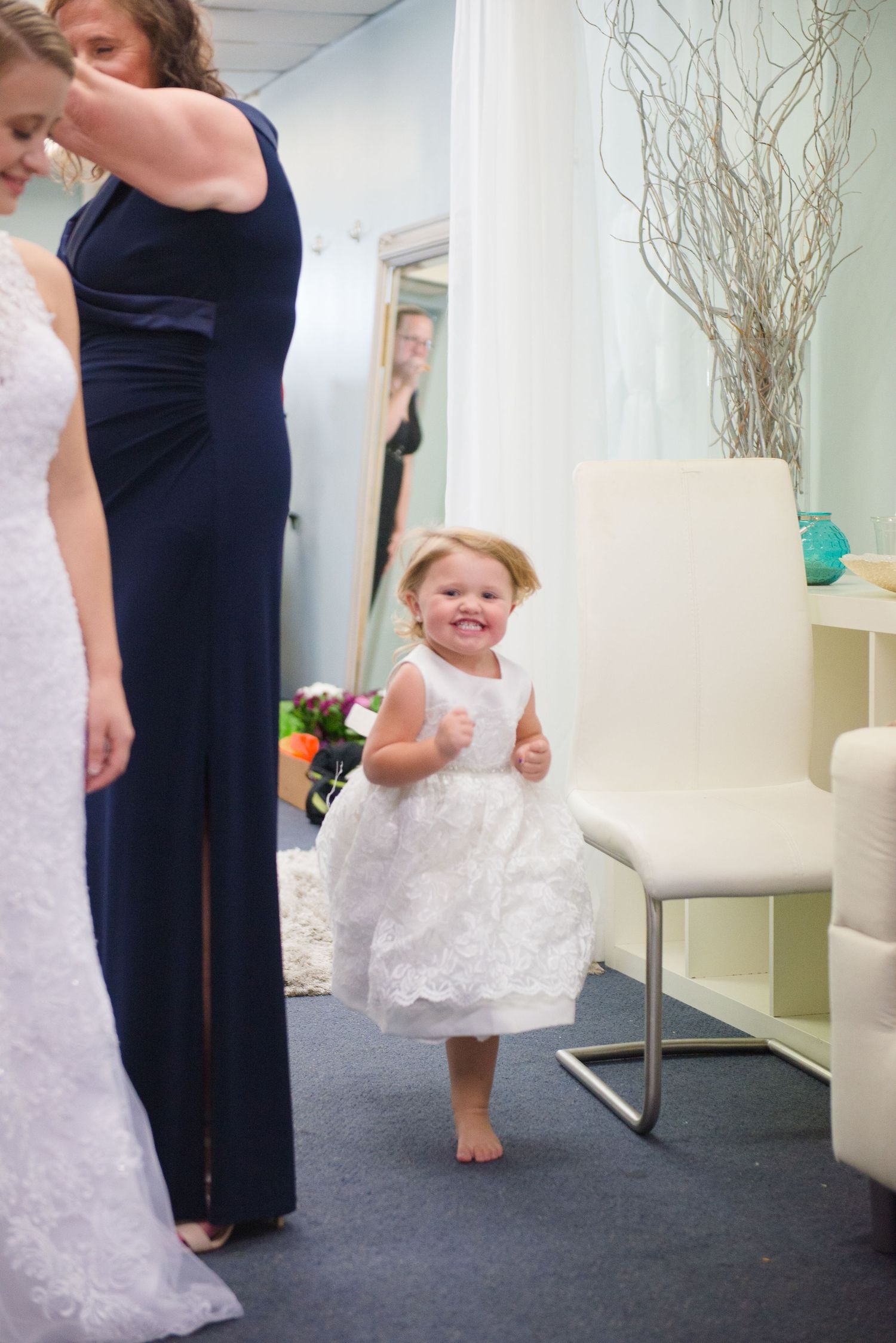 A small child in a white lace dress excitedly watches wedding preparations in a bridal boutique. Andover wedding at Magestic Oaks Golf Club