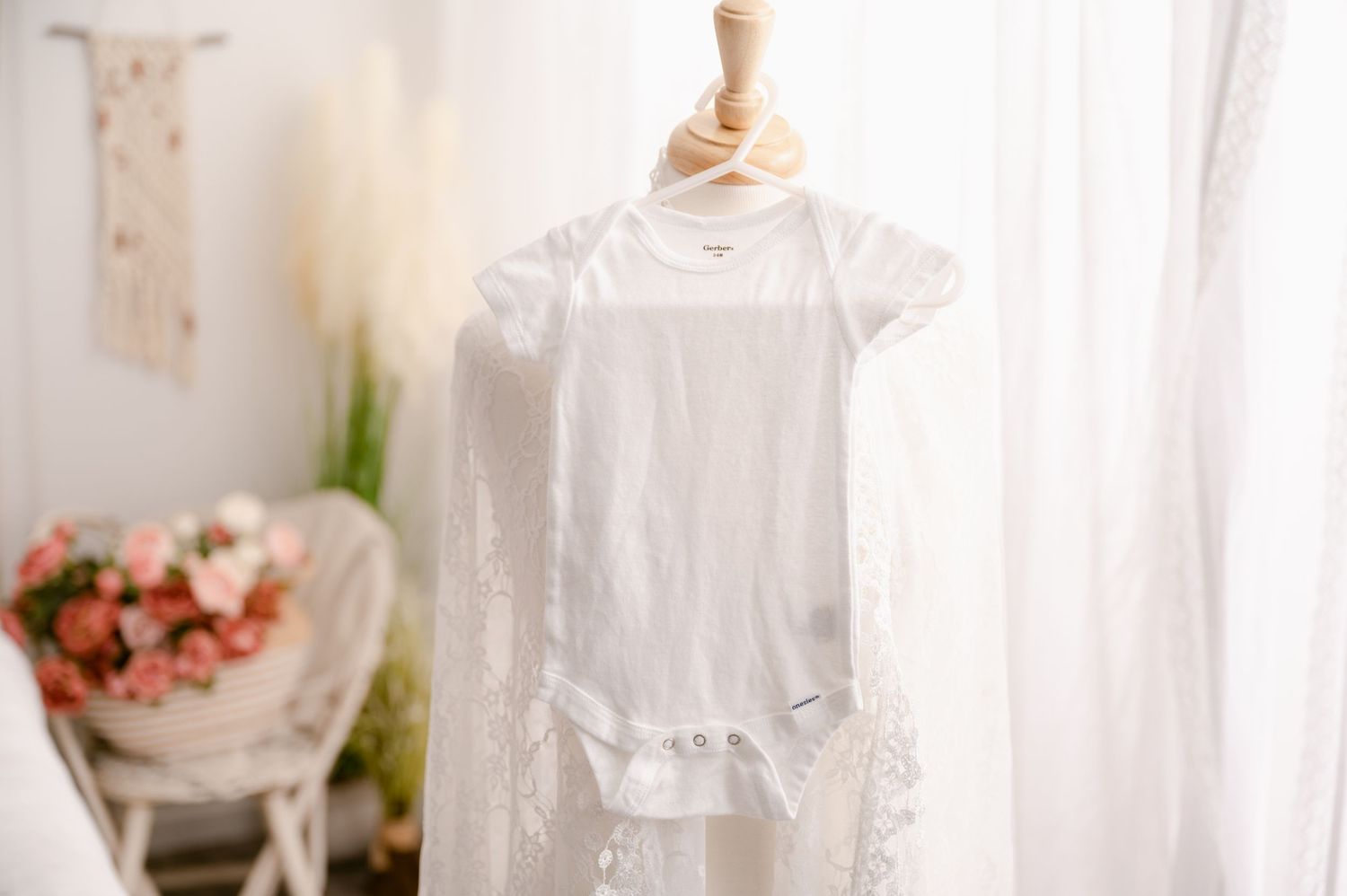 A delicate white christening gown hangs from a cream-colored dress form near sheer curtains and floral decor.