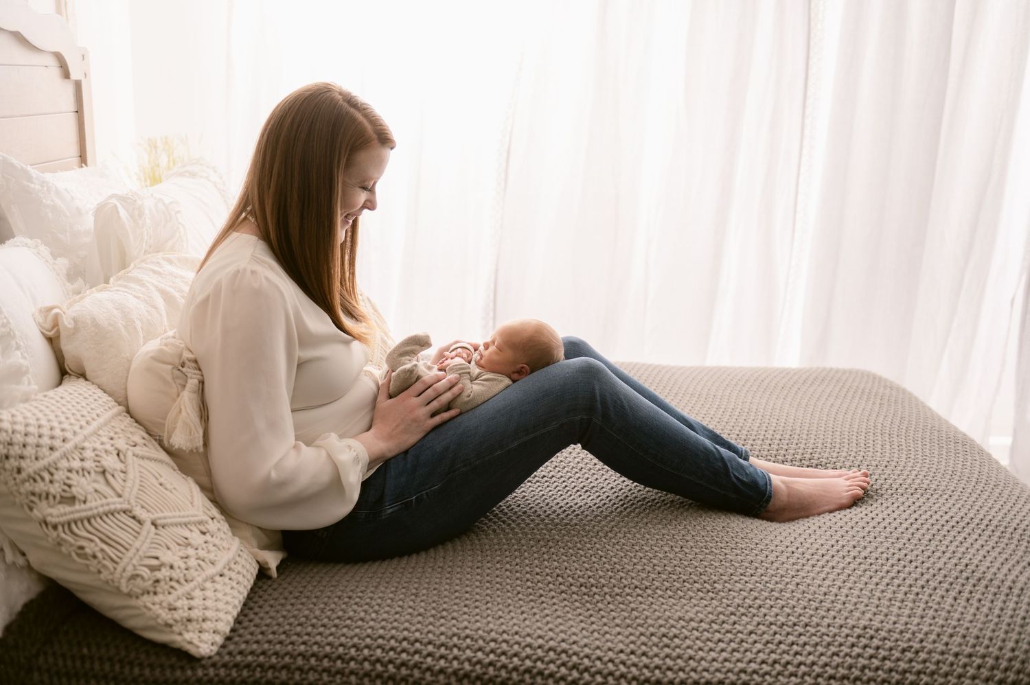 Mother and baby sharing tender moment on grey bed while wearing cozy white sweaters.