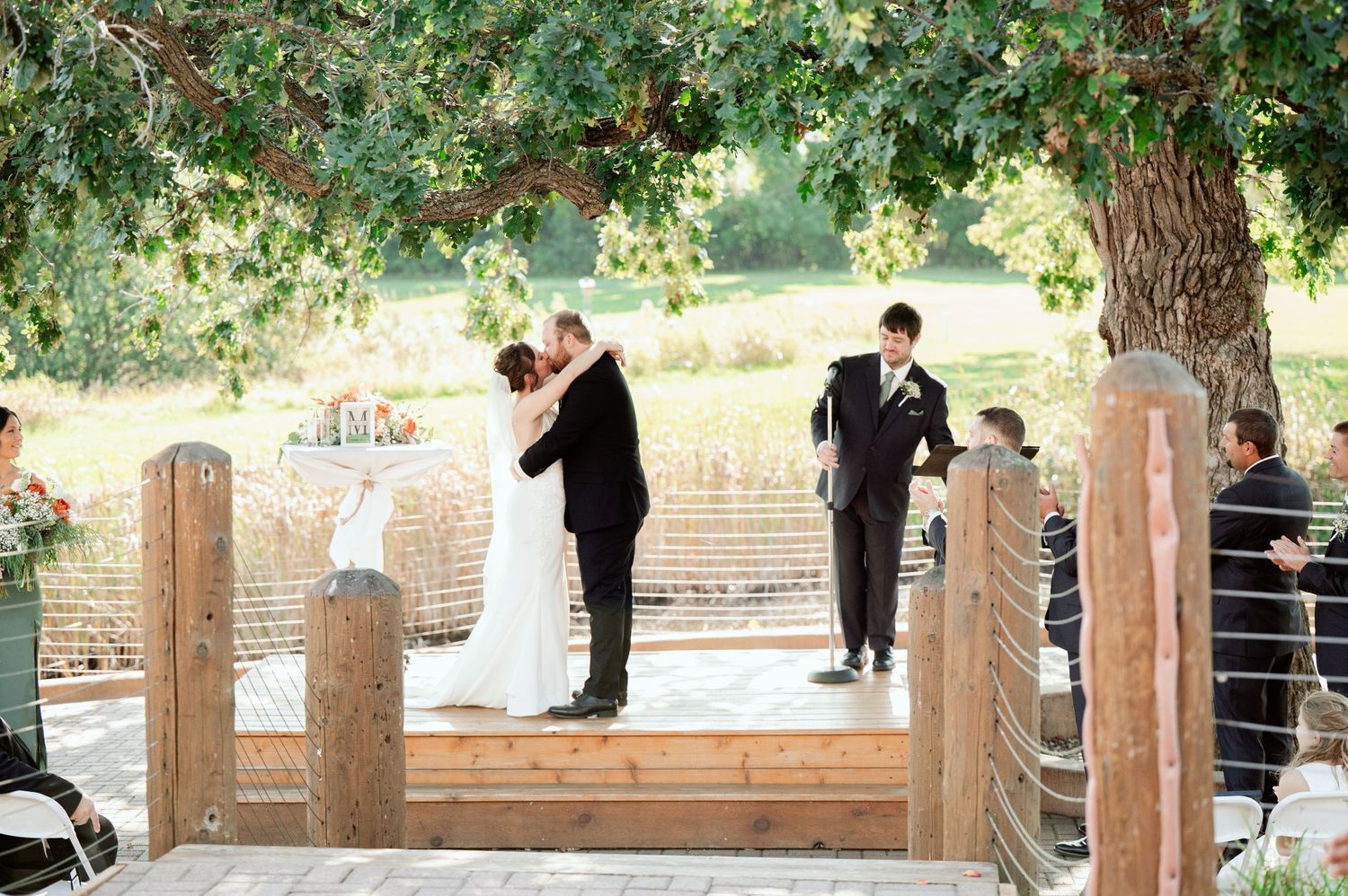 The Grands At Mulligans. A wedding ceremony takes place on a rustic wooden deck surrounded by wooden posts and green foliage.