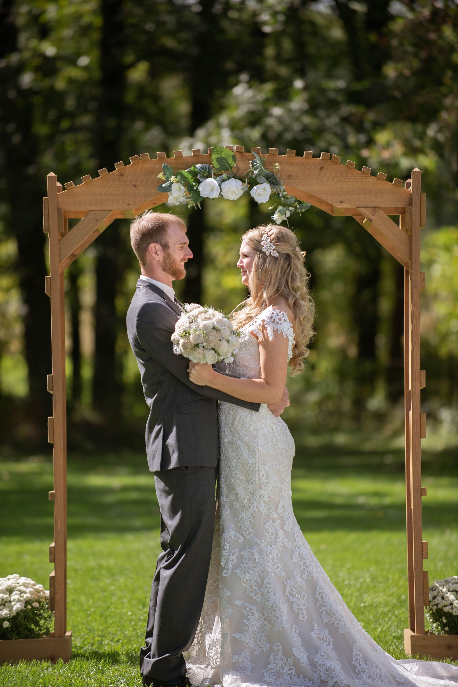 Newlyweds share a romantic moment under a wooden arch decorated with white flowers in a garden setting. Rum River Barn Milaca Minnesota Wedd